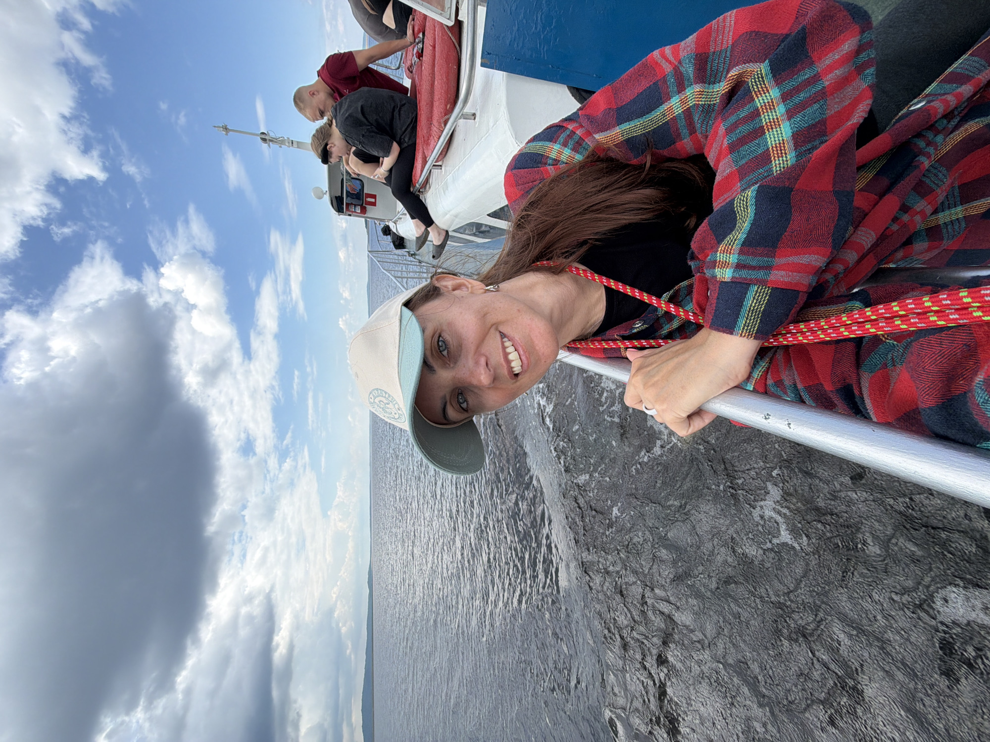 A woman wearing a red and blue plaid shirt and a tan baseball cap smiles as she leans on a metal railing on the deck of a boat. She is looking at the camera. The boat is on a large body of water with a distant shoreline. Other people are visible on the boat in the background, including a man in a red shirt and two younger people sitting down. The sky is cloudy with patches of blue. The location is unknown, Russia.