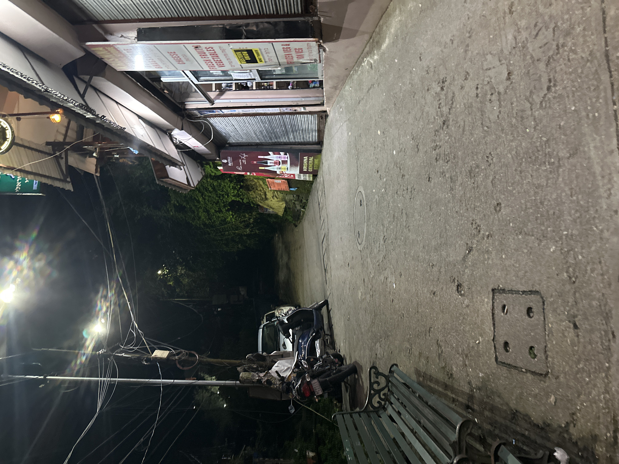 The image depicts a nighttime street scene in an urban environment. A paved road slopes upwards, lined on the right by a storefront with a corrugated metal shutter partially obscuring its entrance. A sign on the storefront reads "KRISHNA GENERAL STORE" and lists "FRESH FRUIT VEGETABLES" and "FROZEN VEG & ION VEG". To the left of the road, a dark green park bench is visible in the foreground. Two motorcycles are parked along the left side of the road. Overhead, numerous electrical wires are strung between poles, illuminated by a bright streetlamp that casts a lens flare. The background is dark, with trees and foliage visible on the left. The ground is concrete with some patches of dirt and debris, and a square metal utility cover with four circular openings is embedded in the pavement.