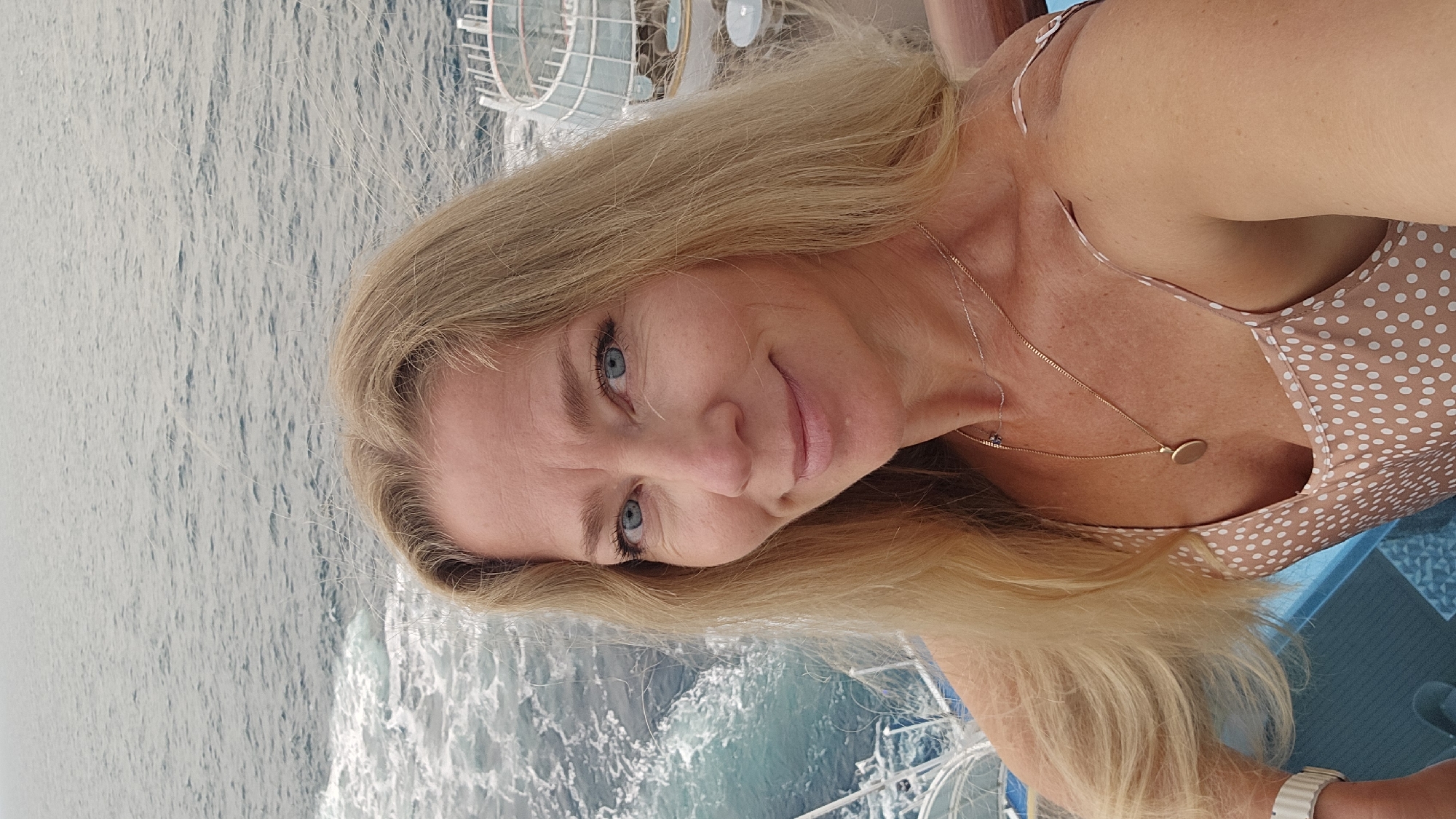 A woman with blonde hair and blue eyes is pictured in a selfie taken on a boat on the sea. She is wearing a polka-dot dress and a gold necklace. The location is assumed to be on the sea, as the boat is moving and the water can be seen. The city location of Unknown, France cannot be confirmed due to the photograph not showing enough identifiable clues.