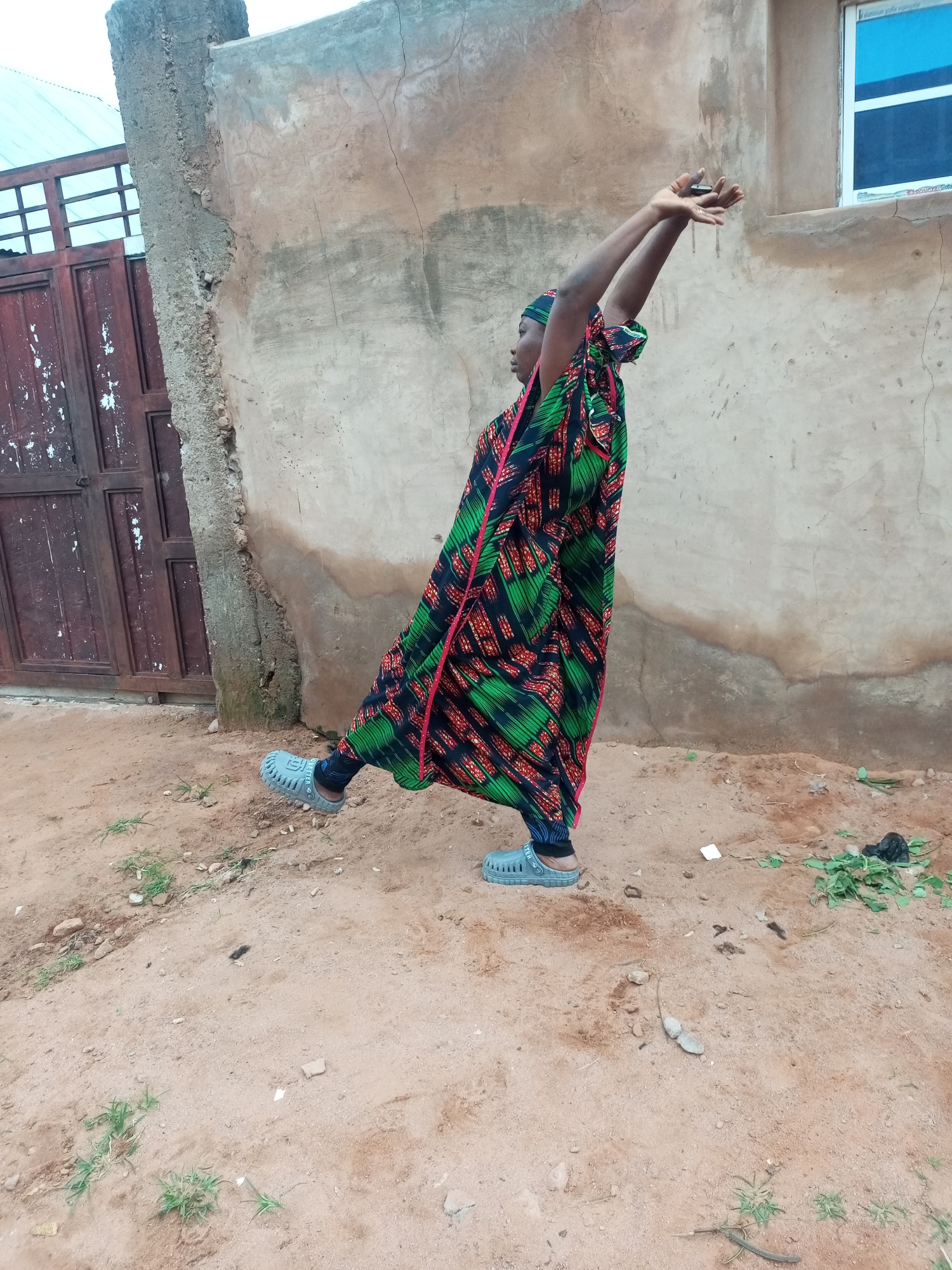 In Tempure, Nigeria, a person stands outdoors on sandy ground in front of a rough, tan-colored building with a single window. The individual is wearing a vibrant, flowing multicolored dress with a pattern of red, green, and black. Their arms are raised above their head, and their hands are clasped together. They are also wearing blue slip-on shoes. To the left of the building, there is a large, weathered wooden gate with metal bars. The ground is uneven and scattered with small stones and sparse green vegetation.