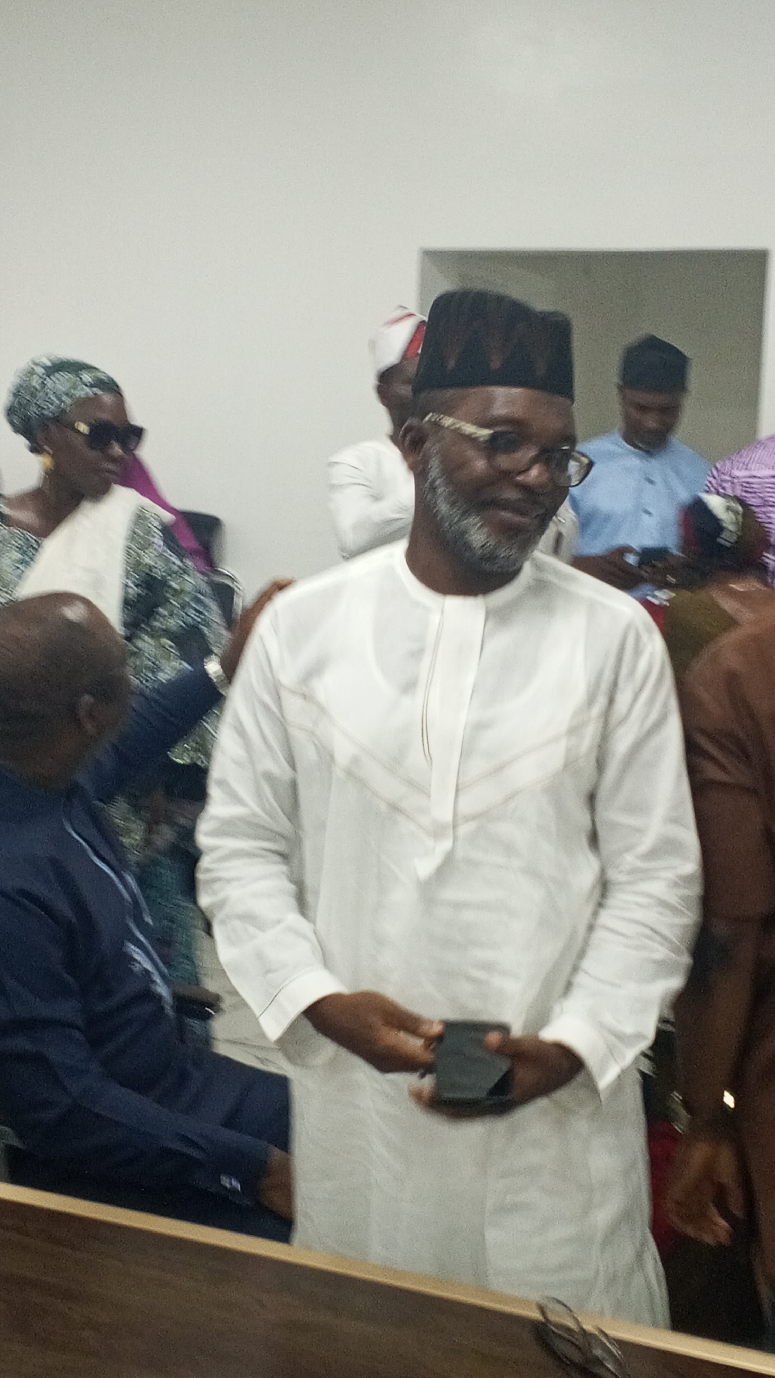 An indoor, eye-level shot captures a gathering of people. The central figure is a man with dark skin, wearing a white long-sleeved tunic with subtle horizontal and V-shaped patterns on the chest. He has a grey beard, wears glasses, and a black, textured cap. He holds a black smartphone in both hands at waist level and looks slightly to his left with a gentle smile.
Around him, several other individuals are visible. To his left, partially obscured, is a woman wearing a patterned headwrap and sunglasses. Another man in a dark blue tunic is seated in the lower-left foreground, mostly obscured. In the background, other people are present; one man wears a light blue tunic, and another has a white and red patterned cap. The setting appears to be a brightly lit room with plain, light-colored walls. A dark wooden surface, possibly a desk or railing, runs across the lower portion of the foreground. The overall atmosphere seems relaxed, consistent with a social event or meeting. No specific time of day or external weather conditions are discernible, and no text is visible in the image.