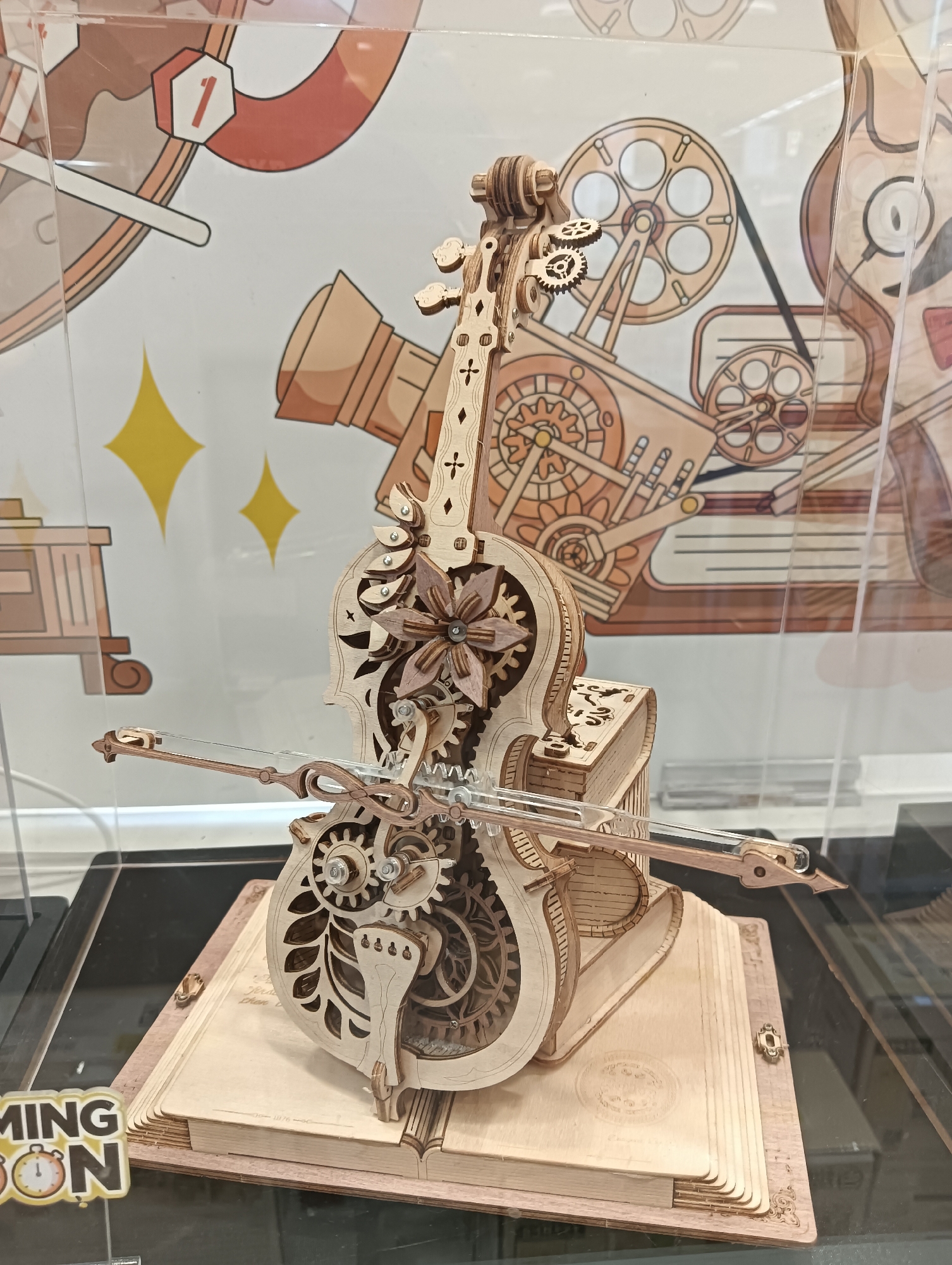 In Kediri City, Indonesia, a detailed wooden model of a violin with intricate gears and a bow is displayed prominently on a wooden book-shaped stand. The violin model, made of light-colored wood, features a decorative floral design on its body and exposed mechanical components that suggest it may be functional. It is positioned on top of an open book, also made of wood, which has decorative etching on its pages and spine. The entire display is enclosed within a clear protective case, likely for preservation and presentation. In the background, a colorful illustration depicts a vintage projector and a stylized character, adding a whimsical and artistic context to the scene. A sign with the text "COMING SOON" and a clock icon is visible in the lower left corner of the image, indicating a promotional or upcoming event. The lighting is bright and even, highlighting the craftsmanship of the wooden model and the surrounding elements.