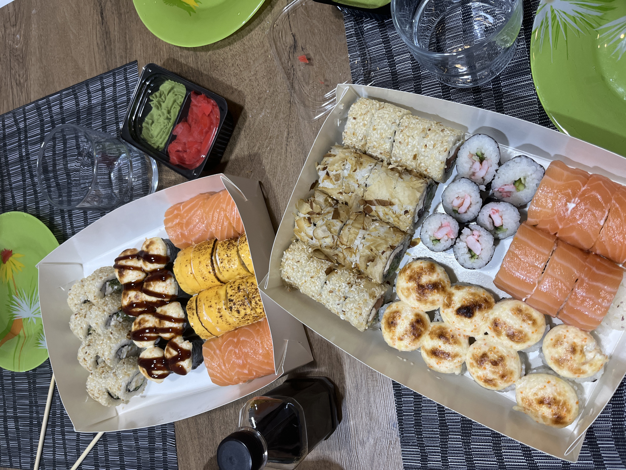 Two open sushi boxes are arranged on a wooden table with a dark striped placemat. The top box contains several pieces of sushi, including rolls covered in sesame seeds, rolls drizzled with a dark sauce, and pieces of salmon. Beside this box, there is a glass of water, a small black container with green wasabi and red pickled ginger, and a bottle of soy sauce. The bottom box holds a variety of sushi, featuring rolls covered in sesame seeds and thinly sliced almonds, small maki rolls with shrimp and cucumber, salmon sashimi, and round, baked sushi pieces with a toasted appearance. A green plate with a floral design is visible in the upper right corner, and another is partially visible in the lower right. The image depicts a meal, likely at a residence in Khmelnytskyi, Ukraine.