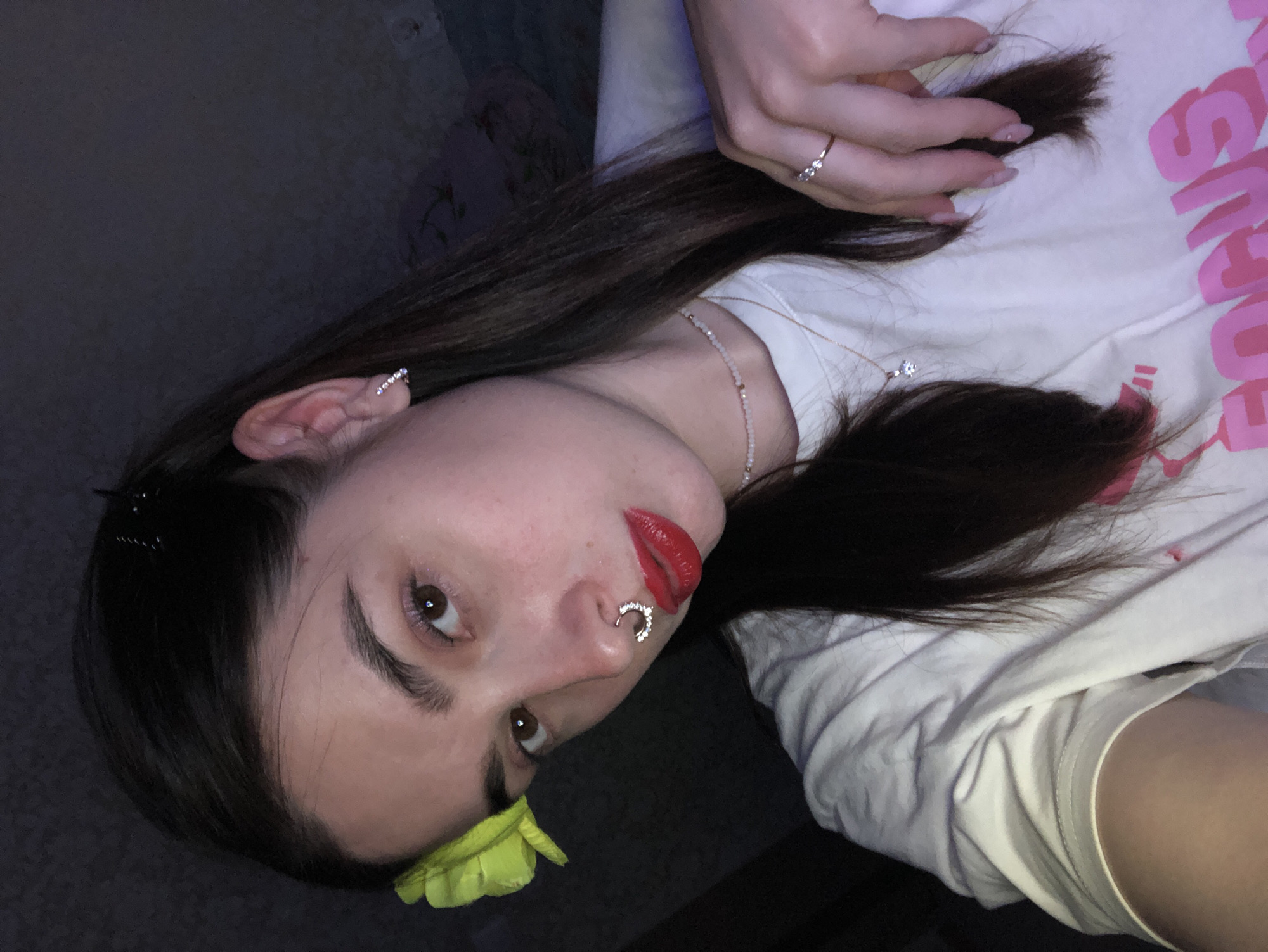 A young woman with long, dark hair and fair skin is the central figure in this selfie. She is wearing red lipstick and has a crescent-shaped nose ring. A yellow flower is placed near her eyebrow. She also has a stud earring and a delicate necklace. Her fingernails are painted with a light pink polish. She is wearing a white t-shirt with pink lettering. Her right hand is holding a strand of her hair. The location is unknown; the description does not include any features indicating Noviy, Russia.