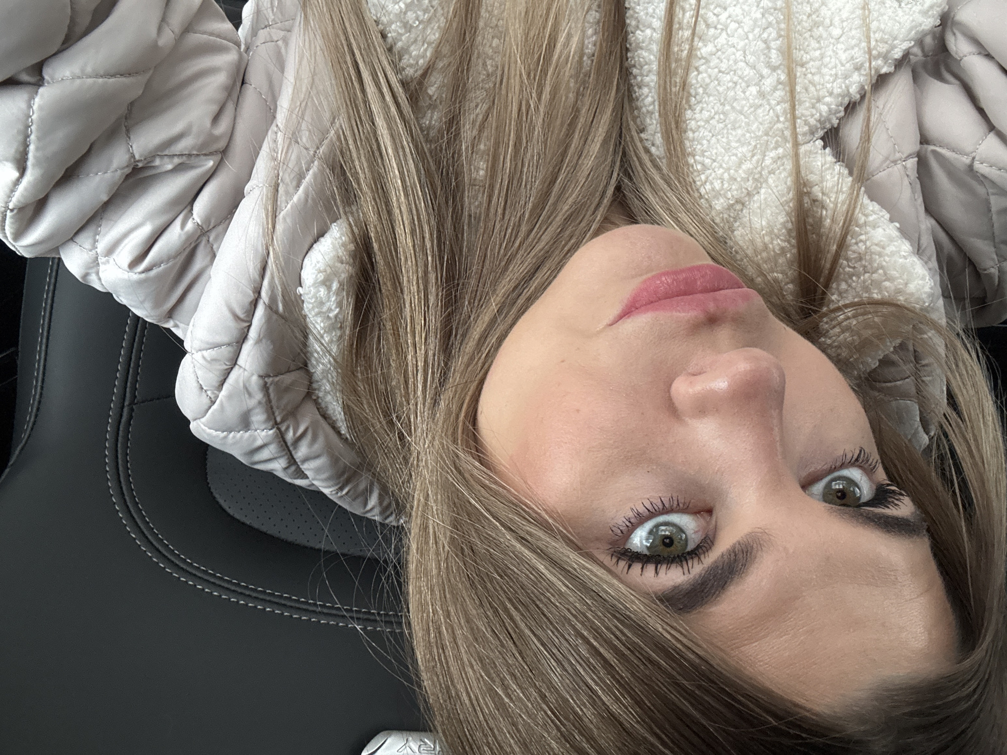 A close-up portrait of a young adult female is depicted, positioned within the interior of a vehicle in Chaikovsky, Russia. The subject's head is tilted, with a direct gaze towards the camera. She has light brown hair, green eyes with visible mascara on both upper and lower lashes, well-defined eyebrows, and pink, slightly parted lips. Her attire includes a light beige quilted jacket, revealing a white, textured, fleece-like lining or collar. The background consists of a dark grey upholstered car seat featuring white stitching and perforated sections. No other individuals or significant actions are visible within the frame.