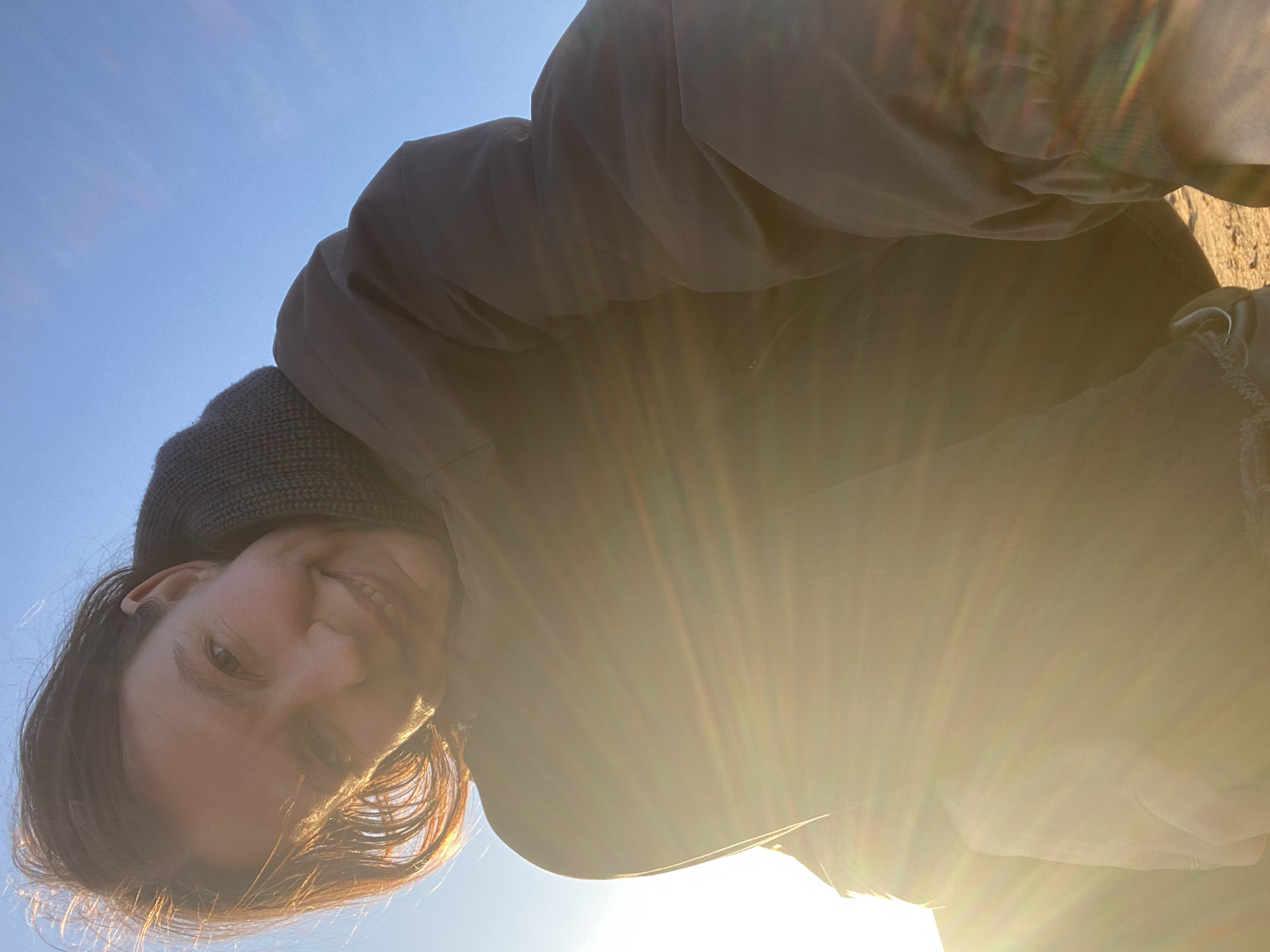 A woman with brown hair and wearing a grey hooded jacket is seen from a low angle, looking down at the camera. The sun is behind her, creating a strong glare and lens flares in the lower portion of the image. The sky is blue in the upper left corner. The setting is outside. There is no information to determine if this image was taken in the city of Chaikovsky, Russia.