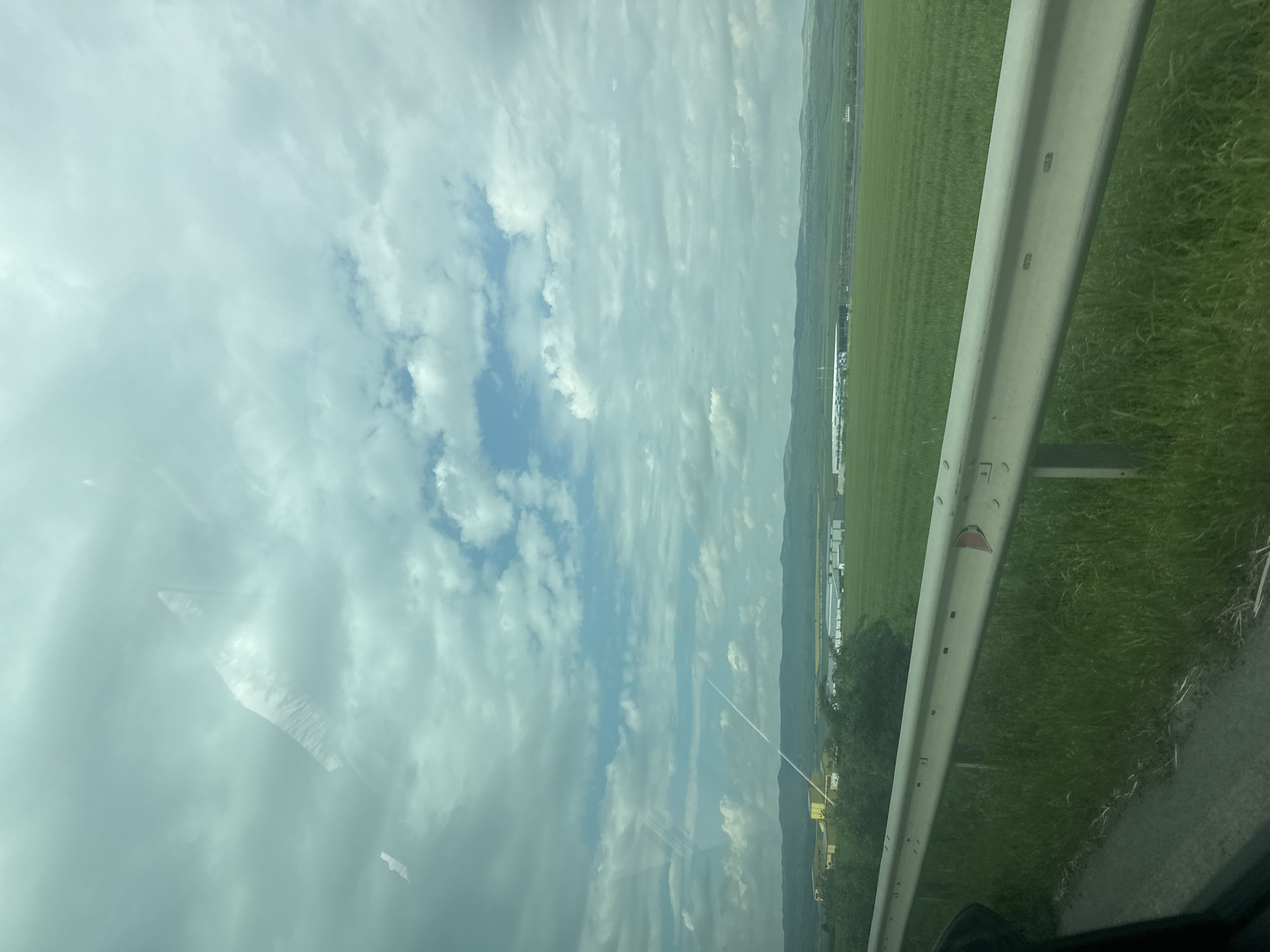 The image depicts a view from a moving vehicle on a road in Burgas, Bulgaria. A metal guardrail is prominently visible on the right, next to a grassy verge. Beyond the verge, a large green field stretches towards a distant, slightly elevated terrain. Several buildings, possibly industrial or agricultural structures, are visible on the field. The sky occupies the majority of the image, showing a mix of clouds with patches of blue sky. The image is slightly blurred, suggesting motion. Reflections are visible on the car's windshield.