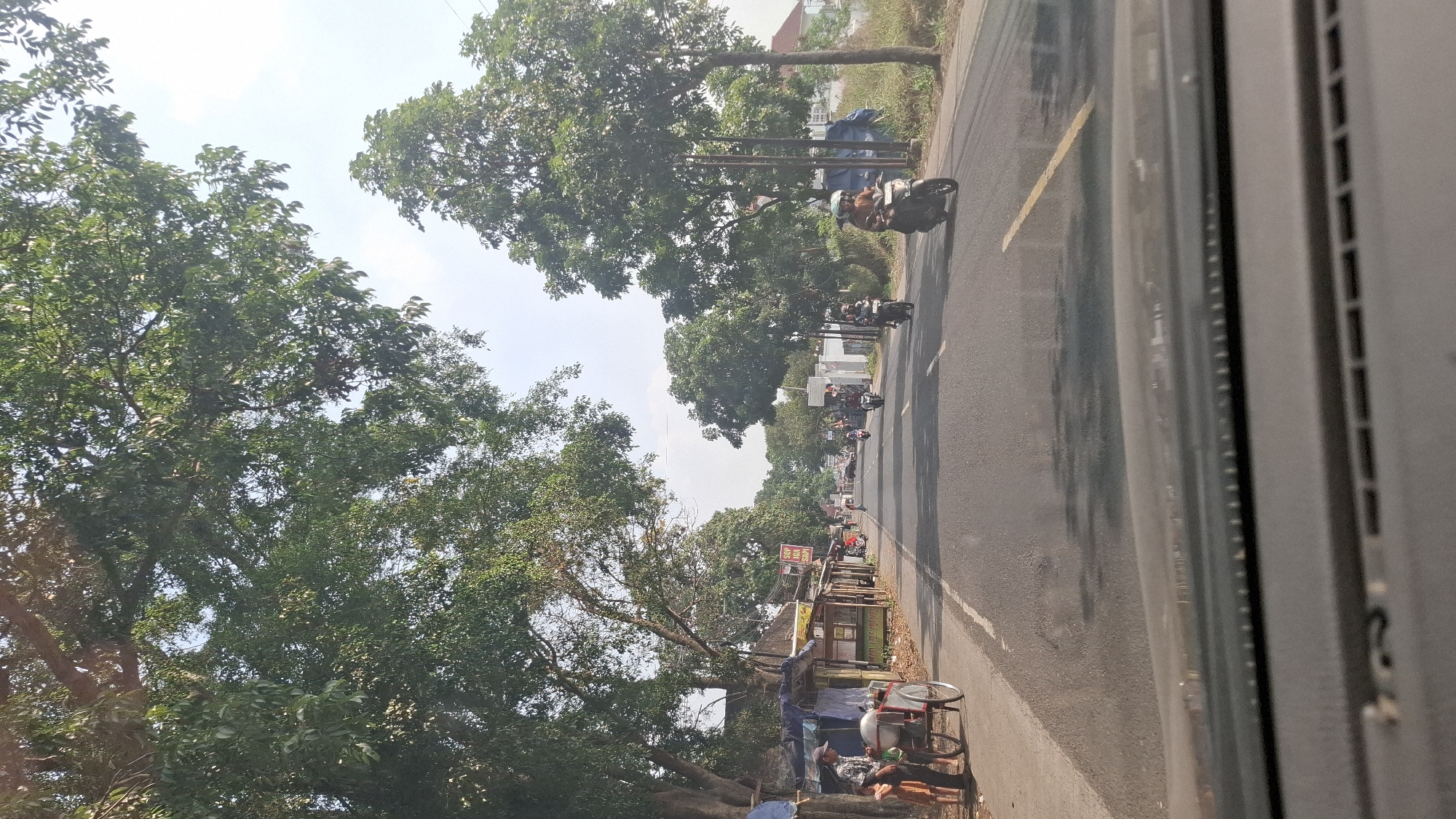 The image depicts a road scene in Warungkondang, Indonesia, likely during the daytime. The road is asphalt, with a yellow line dividing traffic lanes. Several motorcycles are visible, some carrying riders, traveling in both directions. On the left side of the road, there are a few small, simple structures that appear to be stalls or shops, with some people standing near them, including a man interacting with a cart and two children. Trees with lush green foliage line the sides of the road, casting shadows on the pavement. The sky is visible above, appearing bright and possibly indicating a clear or partly cloudy day. The perspective suggests the photo was taken from inside a vehicle.