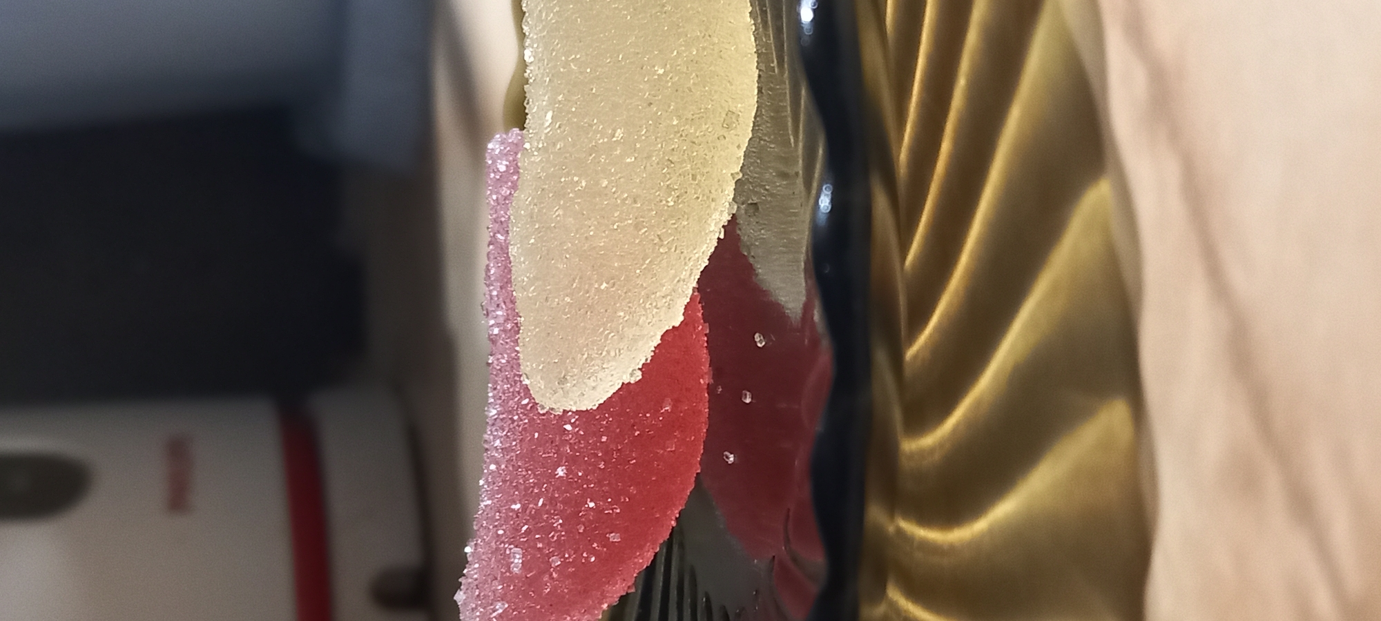 The image is a close-up, primarily featuring two sugar-coated gummy candies on the left side of the frame. The top candy is light yellow or off-white, and the one below it is red or pink. Both are heavily encrusted with sparkling sugar crystals, giving them a textured and lustrous appearance. They appear to be fruit-flavored, possibly shaped like fruit slices.
To the immediate right of the candies, a dark, reflective, and vertically oriented object with a subtle curve is visible, which could be part of a serving utensil or a decorative element. Further to the right, a large section of the image is occupied by draped fabric. The left portion of this fabric is golden, possibly satin or silk, characterized by prominent folds that create a interplay of light and shadow, indicating a soft texture. Adjacent to this, on the far right, is a lighter, peach or beige-colored fabric, also gently folded.
In the softly blurred background on the far left, an indistinct object with a hint of red and white elements can be seen, suggesting a household appliance like a toaster or a kettle. The lighting is bright, illuminating the intricate details of the sugar coating and the sheen of the fabrics. The scene is a still life, and no specific activity is taking place. There is no visible text. The setting appears to be an indoor environment. This image was captured in Kryvyi Rih, Ukraine.