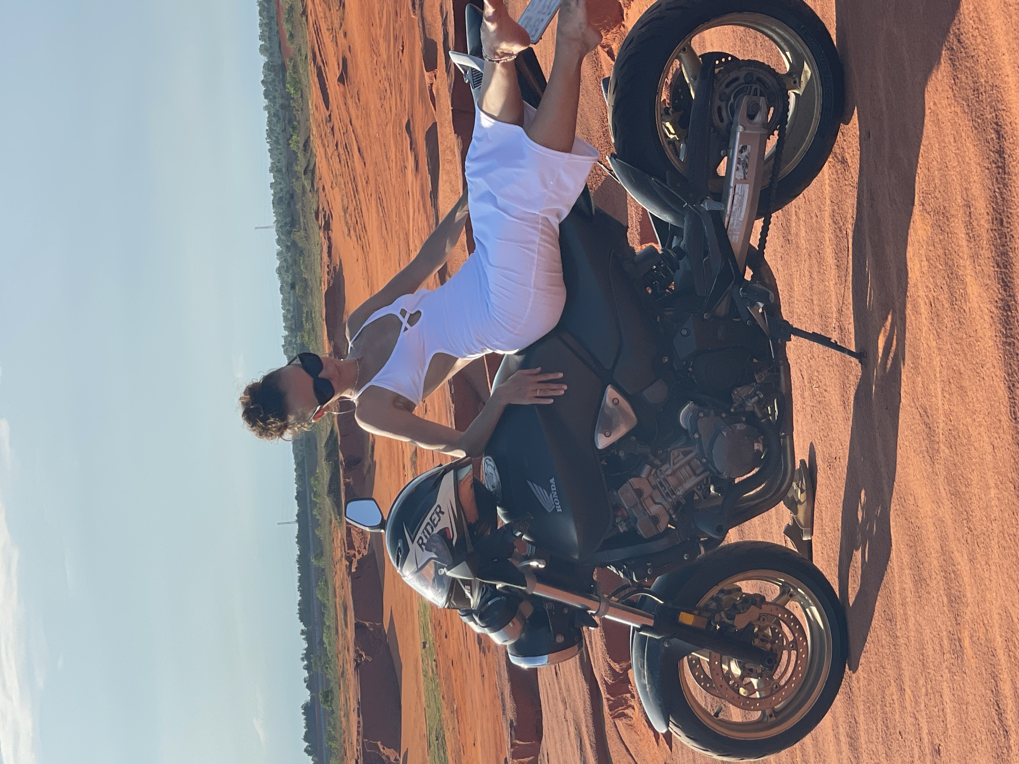 A woman with dark, curly hair pulled up in a bun and wearing sunglasses is posing on a black motorcycle. She is wearing a white, strappy midi dress with cutouts at the sides and chest. Her left leg is extended over the back of the motorcycle, and her right leg is bent. The motorcycle has a helmet with a "Rider" logo on it placed on the handlebars. The scene appears to be set in a desert-like environment with orange sand dunes and a few sparse green trees in the background. The sky is clear and blue. This location is characteristic of the red sand dunes in Mui Ne, Vietnam.