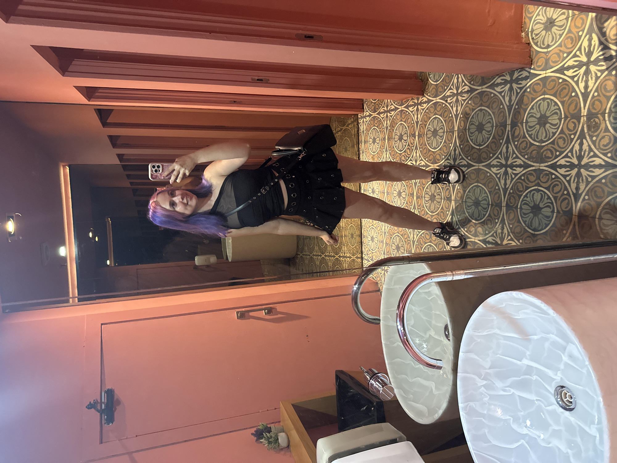 The image shows a young woman with purple and blonde hair taking a mirror selfie in a brightly lit, pink-walled bathroom. She is wearing a black, one-shoulder crop top with mesh detailing and a black mini skirt with grommets. She has a black crossbody bag over her shoulder and is wearing black and white sneakers. The bathroom features ornate, patterned tile flooring, two pink and white marble sinks with chrome faucets, a trash can, and a wooden shelf with small plants. The woman is standing in front of a large mirror that reflects the various stalls and doors of the bathroom. The lighting suggests it is daytime. The style of the bathroom, particularly the tile and pink walls, along with the contemporary decor, could be found in an establishment in Madrid, Spain, possibly a trendy restaurant or bar, though there is no direct evidence to confirm the city.