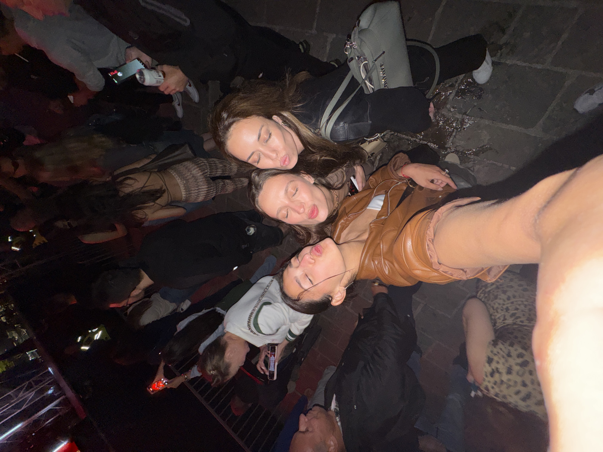 A tilted, high-angle shot shows a group of people, likely in a public space, possibly at night based on the lighting. Three women in the foreground are posing closely together, with their heads touching and lips puckered as if to kiss. One woman is wearing a brown leather jacket, and another a white sweatshirt. People with various attire and some holding beverages and mobile phones surround them. A green bag is visible next to the women. The ground appears to be stone and somewhat wet. The location is unknown, and does not appear to be in Budva, Montenegro.
