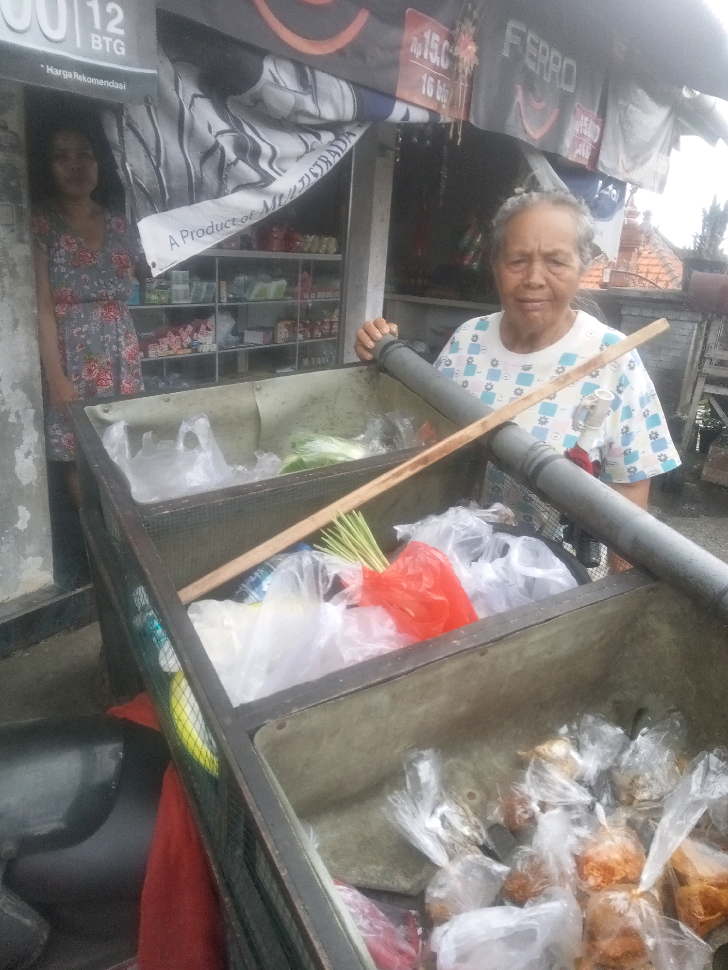An elderly Indonesian woman stands behind a market cart filled with plastic bags of produce and packaged goods, in what appears to be the city of Mendoyo, Indonesia. She is wearing a white t-shirt with a blue and green floral pattern. The cart is made of metal and wood, with a mesh covering. In the foreground, plastic bags contain various items, including what looks like lemongrass and an orange bag with red contents. Behind the woman, shelves are stocked with products, and a sign overhead reads "FERRO." A young woman in a floral dress is partially visible to the left of the cart. The background suggests an outdoor market setting, with a tiled roof structure visible in the distance.