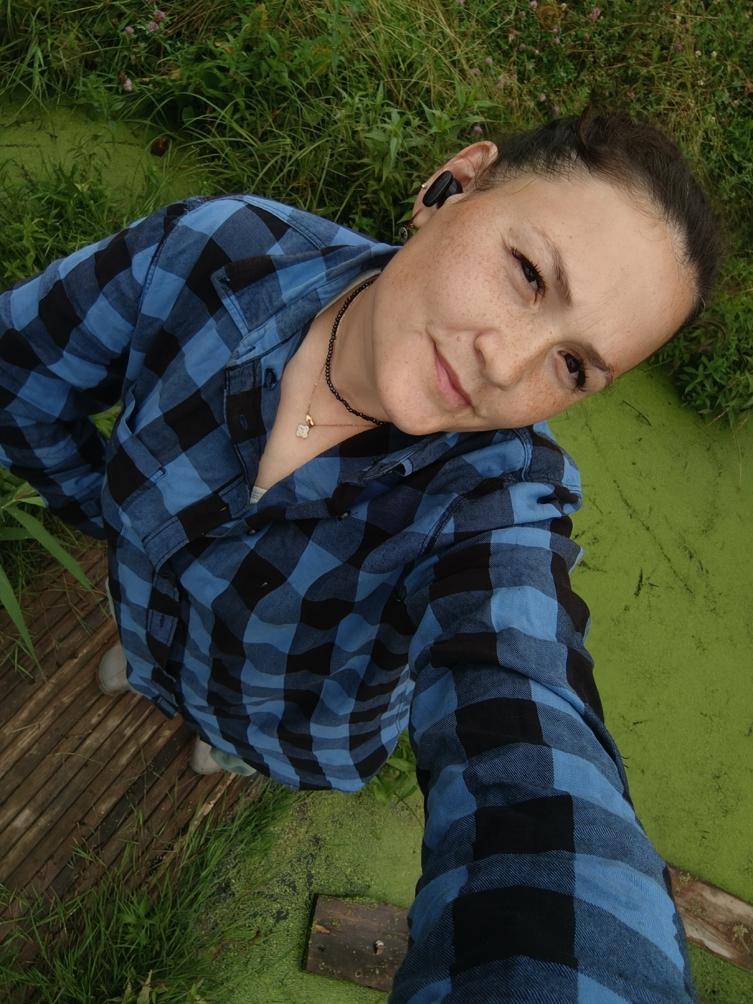 A high-angle, full body selfie shows a woman with freckles standing on a wooden path next to a body of green water. She is wearing a blue and black checkered shirt, and her hair is pulled back. She is wearing a black necklace with a small, gold clover pendant. She has a black earbud in her right ear. The area appears to be a natural outdoor setting, possibly a park or wetland. The image was taken in the city of Karsha, Russia.
