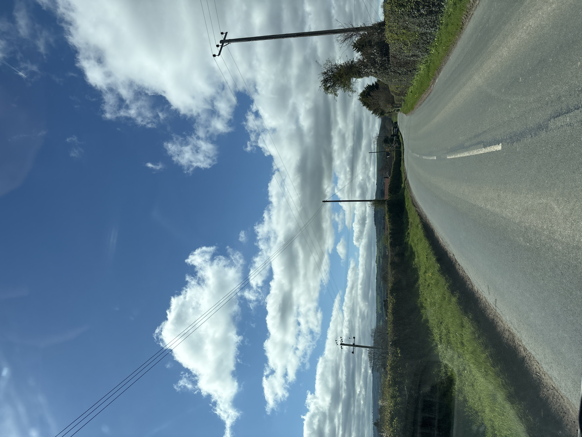 The image shows a rural road in Longden, United Kingdom. The road is asphalt, with a broken white line marking the center. Verges of grass and hedgerows line both sides of the road. Overhead power lines are visible, supported by wooden poles. The sky is blue with scattered white clouds. No people are visible.