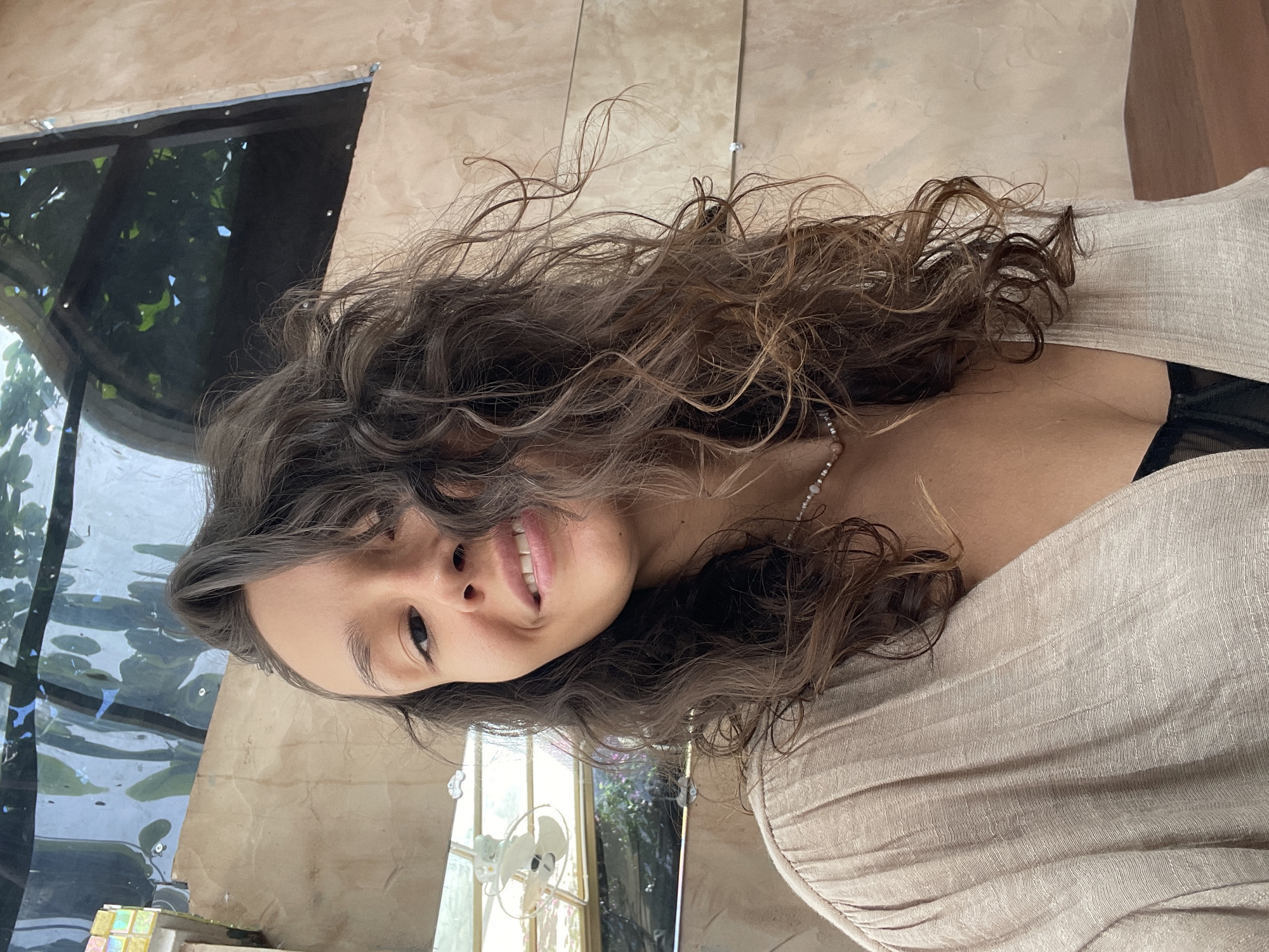 The image features a smiling young woman captured in a medium-close portrait, looking directly at the camera. Her long, dark brown, curly hair cascades around her shoulders and frames her face. She has dark eyes and a cheerful expression, with a slight open-mouthed smile revealing her teeth. She is wearing a light beige or off-white top made of a textured fabric, possibly linen, with a black undergarment visible at the neckline. A delicate, light-colored beaded necklace is visible around her neck.
The setting appears to be indoors, likely near a large window or opening that offers a view of a lush outdoor environment, consistent with the location of Mũi Né, Vietnam. To her left, behind her head, is a dark-framed window showcasing green tropical foliage and a body of water, possibly a pond or pool, reflecting light. To her right, a light brown tiled wall is visible, along with a glimpse of another window in the distance where a small white fan can be seen, surrounded by more greenery. The lighting is bright and natural, indicating it is daytime, and the overall mood is relaxed and pleasant. No text is visible in the image.