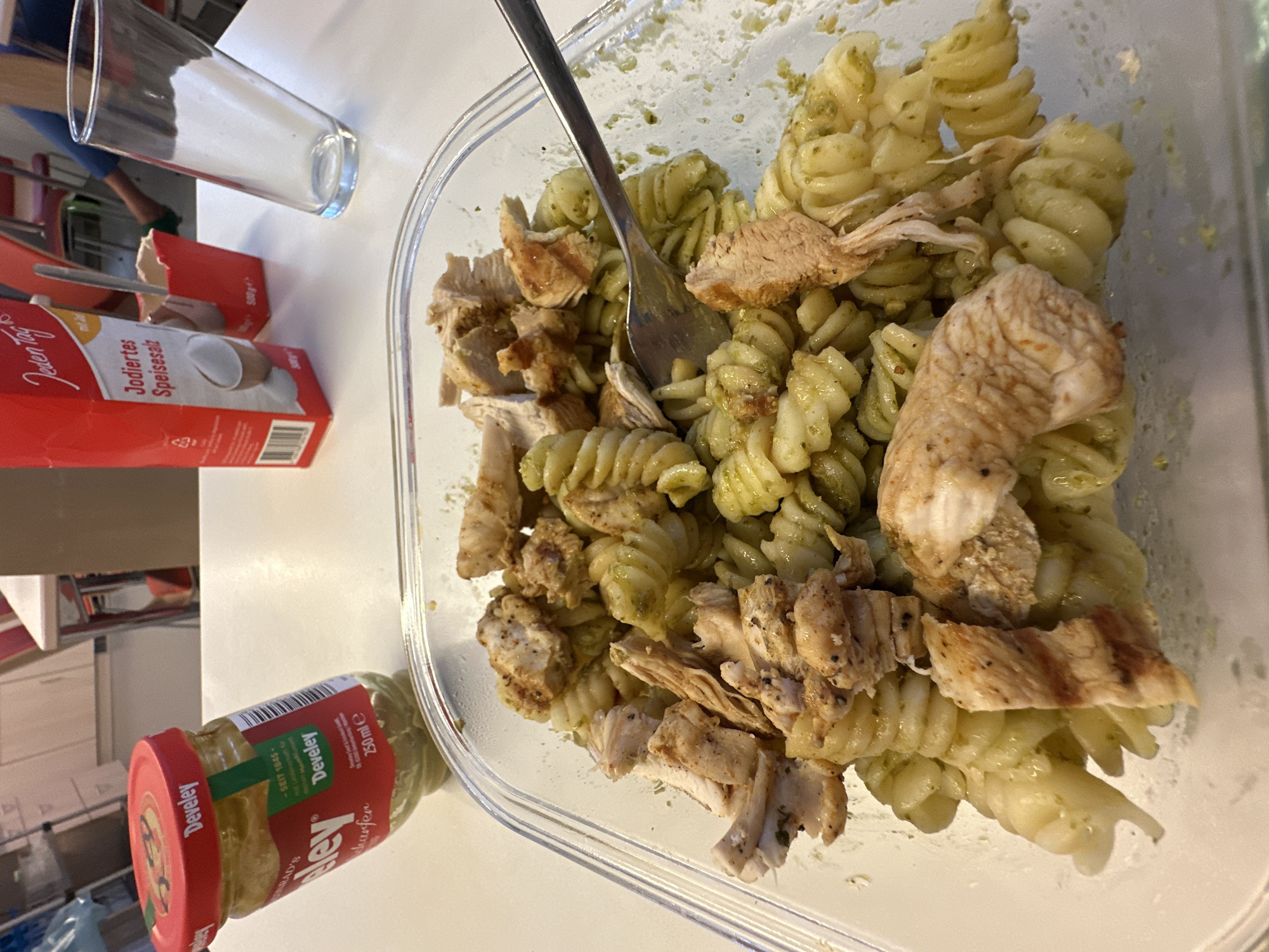 The image shows a close-up view of a meal consisting of rotini pasta with a green sauce, topped with sliced chicken breast. The meal is served in a clear plastic container. To the left of the container is a jar of Develey mustard. In the background, a box of "Jodiertes Speisesalz" (iodized table salt) from "Jeden Tag" is visible, along with a glass of water and a small red paper container holding a spoon. The setting appears to be a table in what could be a kitchen or dining area, with hints of furniture and shelving in the background. The lighting suggests an indoor environment. There is no indication of the city of Garmisch-Partenkirchen, Germany, in the image or its context.