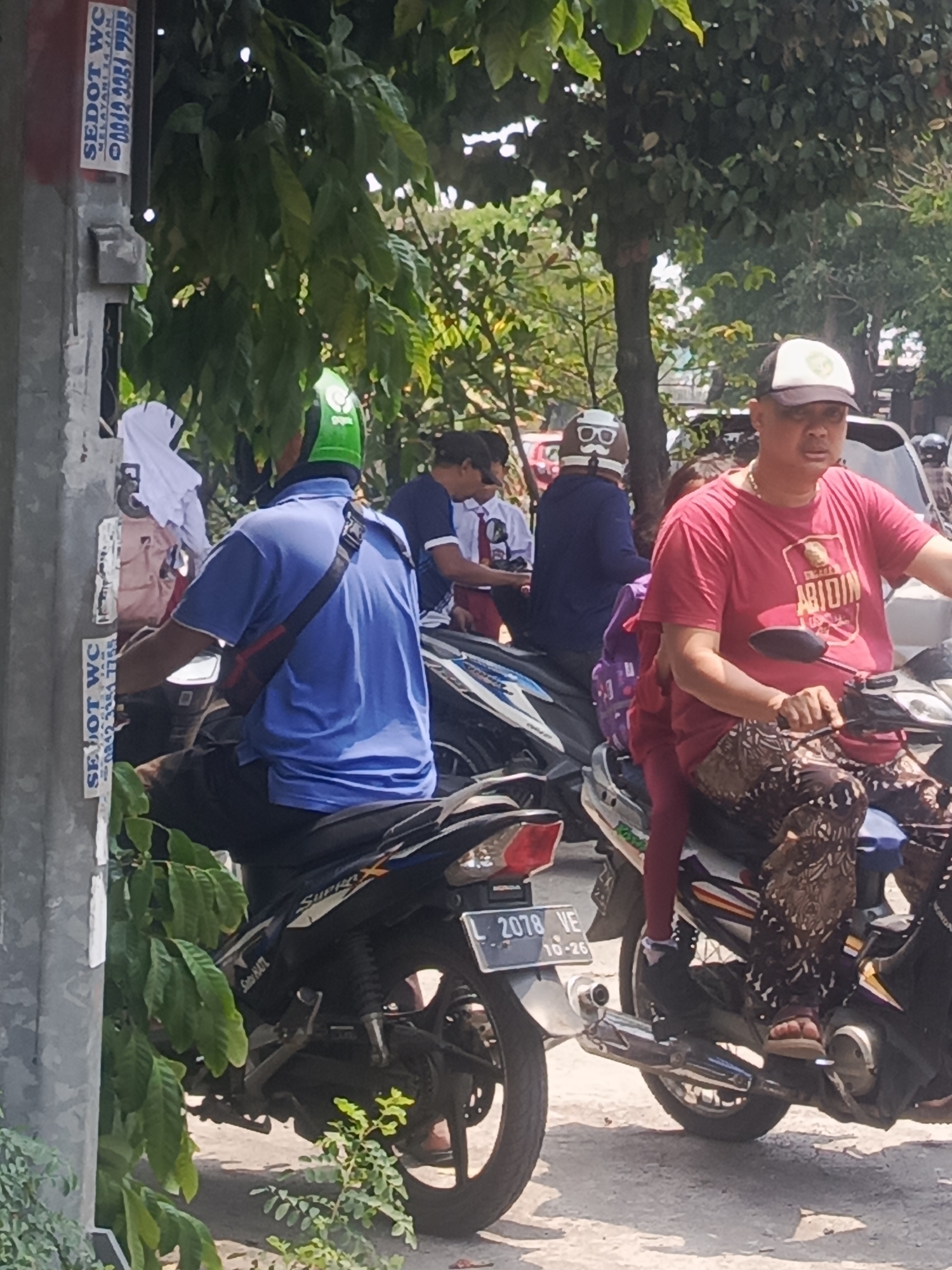 In Surabaya, Indonesia, two men on motorcycles are seen from the side. The man in the foreground wears a red t-shirt with the word "ABIDIN" on it, a black baseball cap, and patterned pants. He is riding a black motorcycle and has a younger person, possibly a child, riding behind him, wearing a purple backpack and a pink outfit. To the left, another man is wearing a blue shirt and a green helmet, also riding a motorcycle. In the background, other individuals and vehicles are partially visible, suggesting a street scene with moderate activity. A sign with text in Indonesian and a phone number is affixed to a pole on the left side of the image. The environment appears to be outdoors, with trees and foliage providing shade.