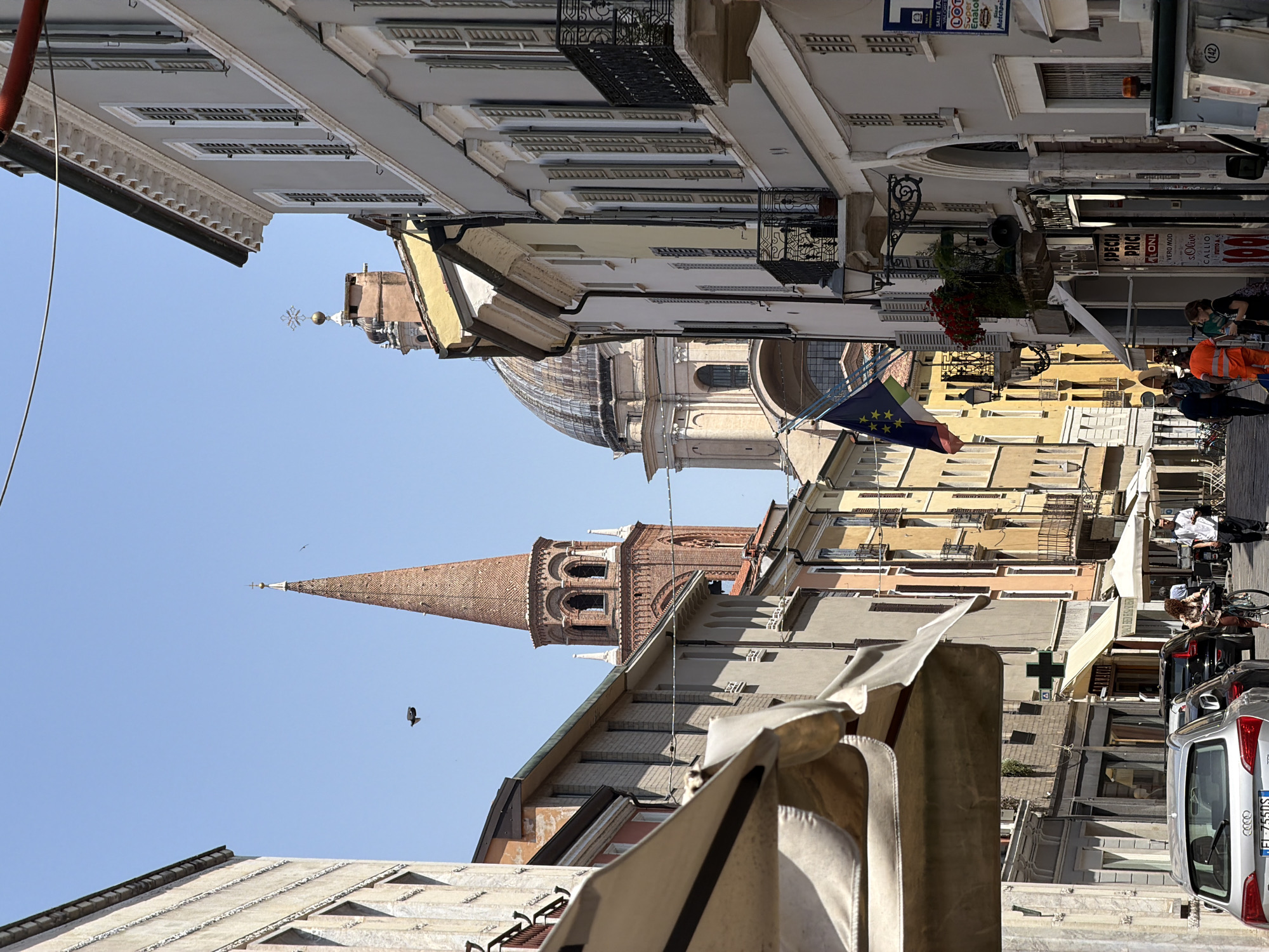 The image depicts a street scene in Mantua, Italy, characterized by buildings of varying heights and architectural styles, indicative of a historic urban environment. In the center of the frame, a tall brick spire with arched windows and a pointed roof dominates the skyline, slightly off-center to the left. Adjacent to the spire is a dome-shaped structure, possibly a church or cathedral, which gives a sense of the city's religious and historical significance.
The street is lined with buildings, some of which have awnings extending over the sidewalks. There is an Italian flag visible among the buildings. The image includes people walking, and parked vehicles along the street.