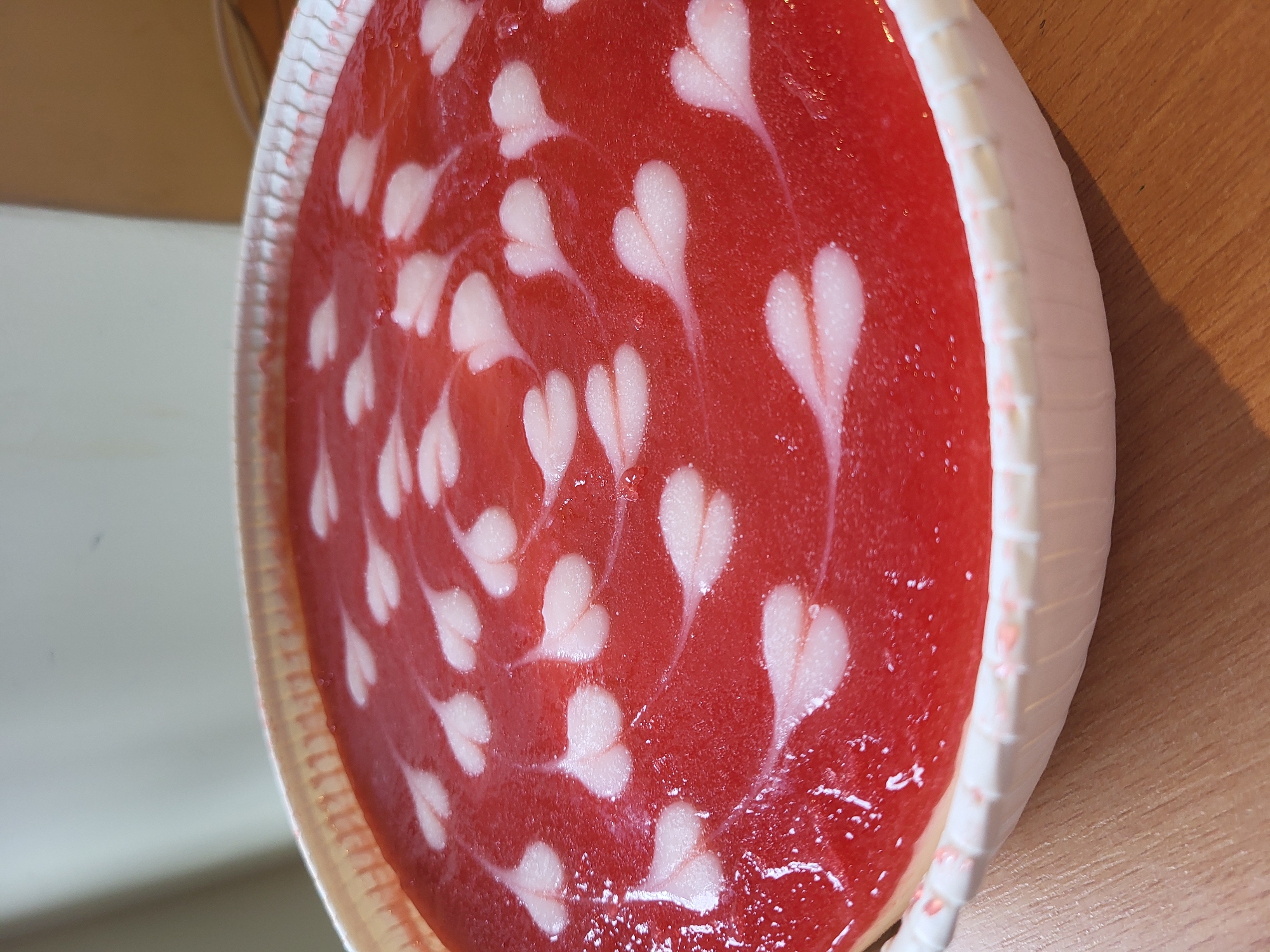 A close-up, high-angle shot shows a round, red dessert in a white fluted paper liner, sitting on a wooden surface. The dessert has a glossy surface with a swirling pattern of small, white heart shapes. The hearts are arranged in concentric circles, with some lines of white liquid connecting them. The dessert appears to be a cheesecake or a jelly-like confection. The background is blurred, with a light-colored vertical surface on the left and a darker, angled surface on the right, suggesting an indoor setting. The lighting is soft and even. The scene is devoid of people or any other objects.