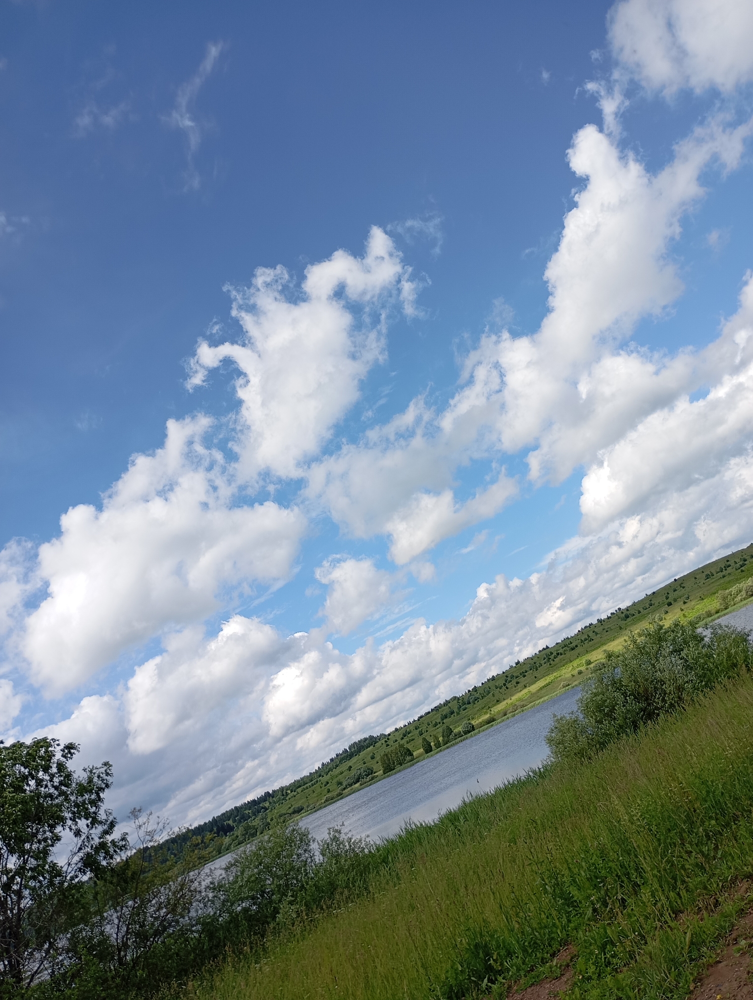 The image depicts a scenic natural landscape in the village of Ryabki, Russia, under a vast, partly cloudy blue sky. In the foreground, lush green grass and bushes dominate the scene, with a dirt path visible in the lower right corner. A tranquil body of water, likely a river or lake, stretches across the middle ground, reflecting the sky. The far bank of the water is lined with rolling green hills dotted with trees, creating a picturesque backdrop. The overall impression is one of a peaceful and serene rural environment. There are no people or animals visible in the image.