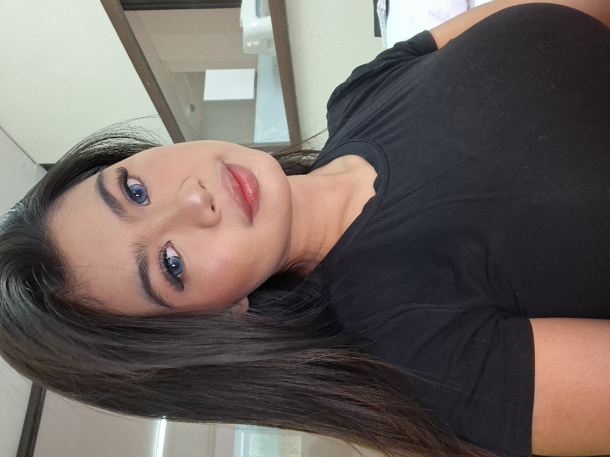 A medium close-up selfie shows a young woman with dark hair, fair skin, and blue contact lenses. She is wearing a black t-shirt and is looking towards the camera with a slight smile. Her makeup is noticeable, with emphasis on her eyes and lips. The background appears to be an indoor setting, possibly a room in Kuta, Indonesia, with shelves and some objects visible. The lighting is bright, suggesting it is daytime.