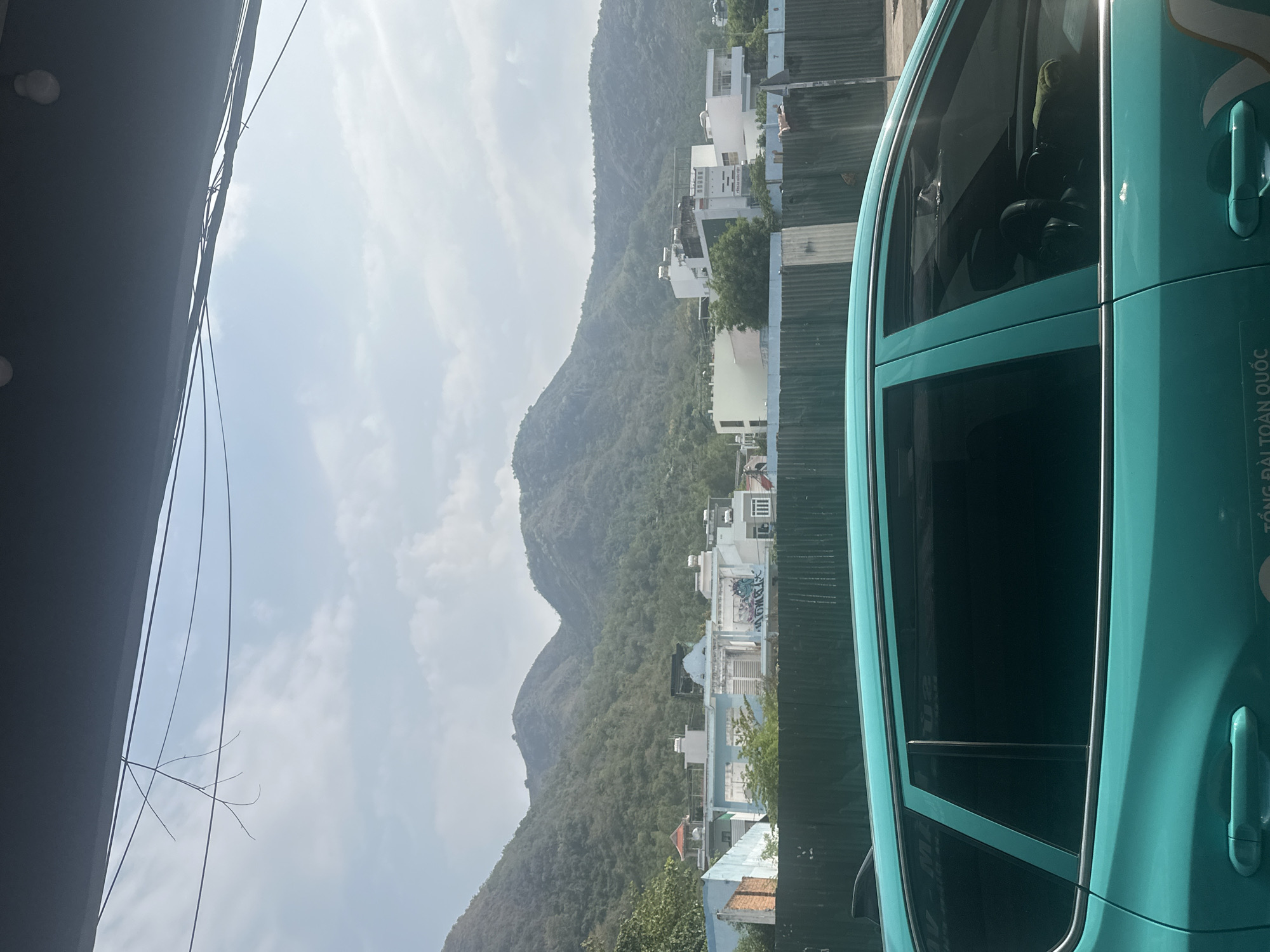The image shows a partial view from inside a turquoise car, with the backdrop of Bac Nha Trang Ward in Vietnam. A mountain range is visible through the car window, dotted with white buildings and lush greenery. The sky is overcast with some glimpses of sunlight. Details of the car's interior, like the seats, are visible in the reflection of the window. A metallic green fence can be seen to the right of the houses. A building awning and a mess of wires are visible on the left.