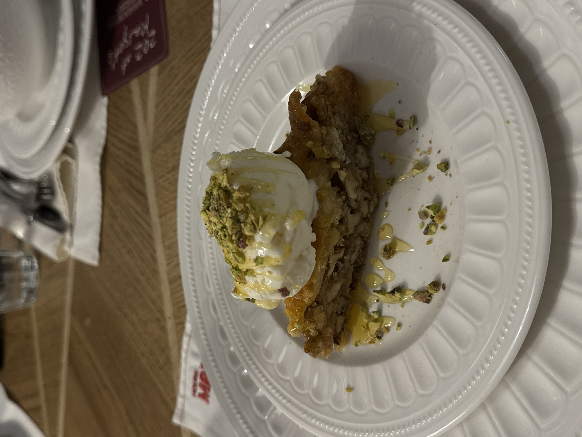A bird's eye-view depicts a plate of pistachio-topped baklava, drizzled with syrup, and a scoop of vanilla ice cream. The plate has a decorative, beaded edge. The image was likely captured at a restaurant in Yerevan, Armenia, given the branding on the napkin visible. The table appears to be made of wood. A stack of plates, a glass, and a menu or bill are visible on the left.