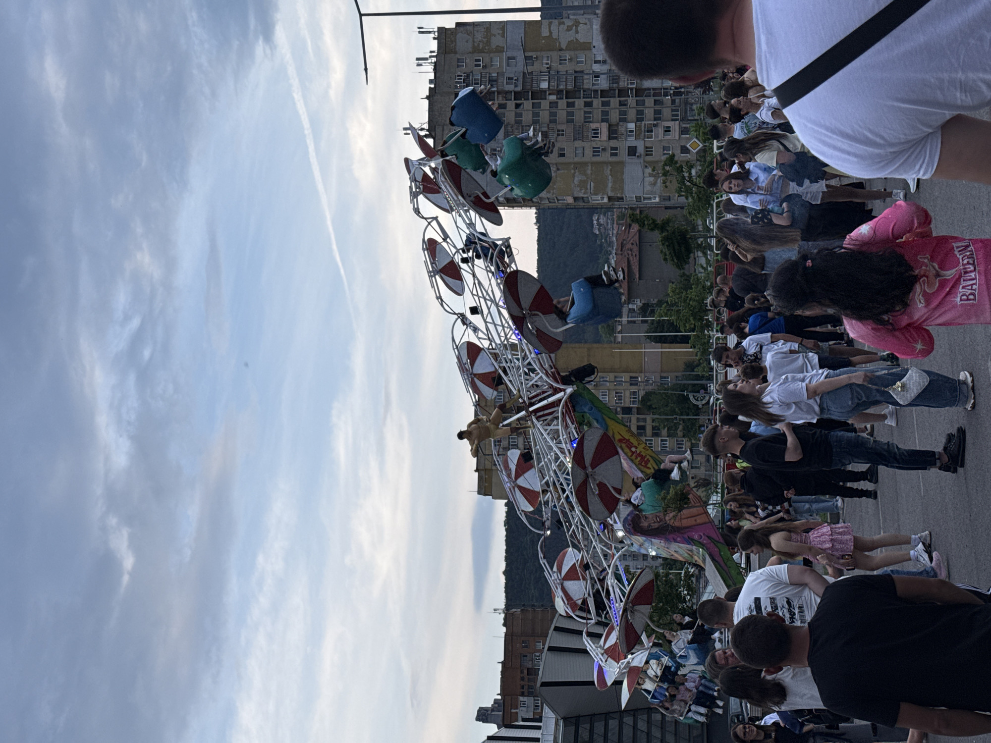 The image shows a rotating amusement park ride in the city of Shumen, Bulgaria. The ride has several buckets with a red and white awning on top. People are riding in the buckets and observing. A crowd of people are present, including a person wearing a pink jacket. In the background, there is an apartment complex and mountain. The sky is mostly cloudy.