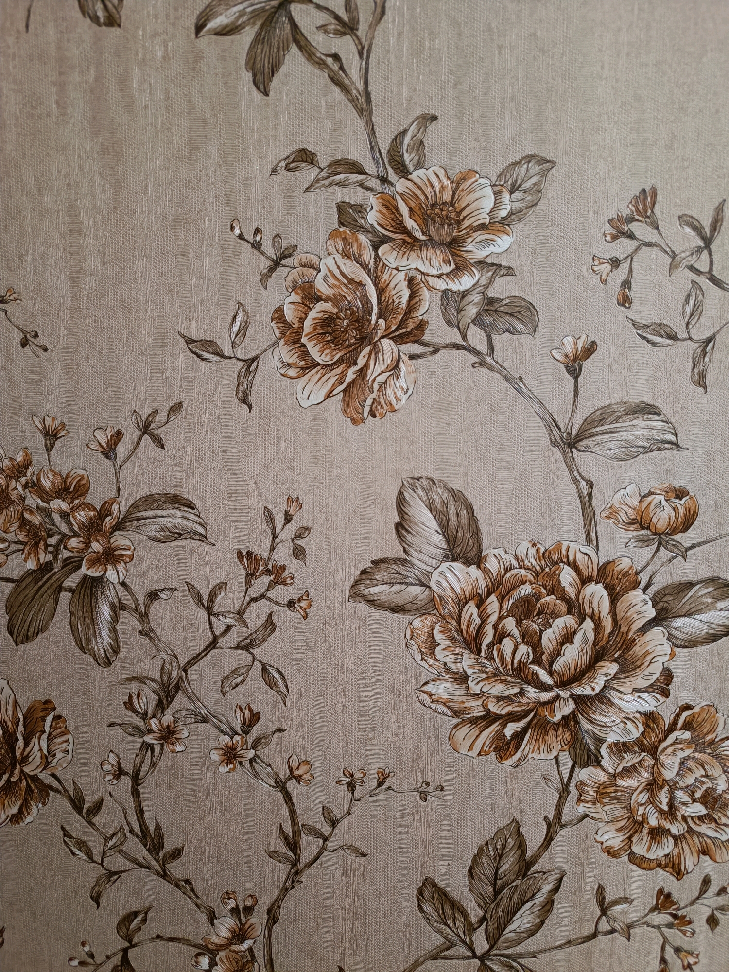 This image is a close-up, top-down, eye-level, medium shot of a beige textured wallpaper with a brown floral pattern. The pattern features large, detailed flowers with many petals, as well as smaller buds and leaves, all rendered in shades of brown and white. The wallpaper has a subtle, embossed texture. The image does not depict any people, objects, actions, or interactions, nor does it provide any information about a location in Srinagar, India.