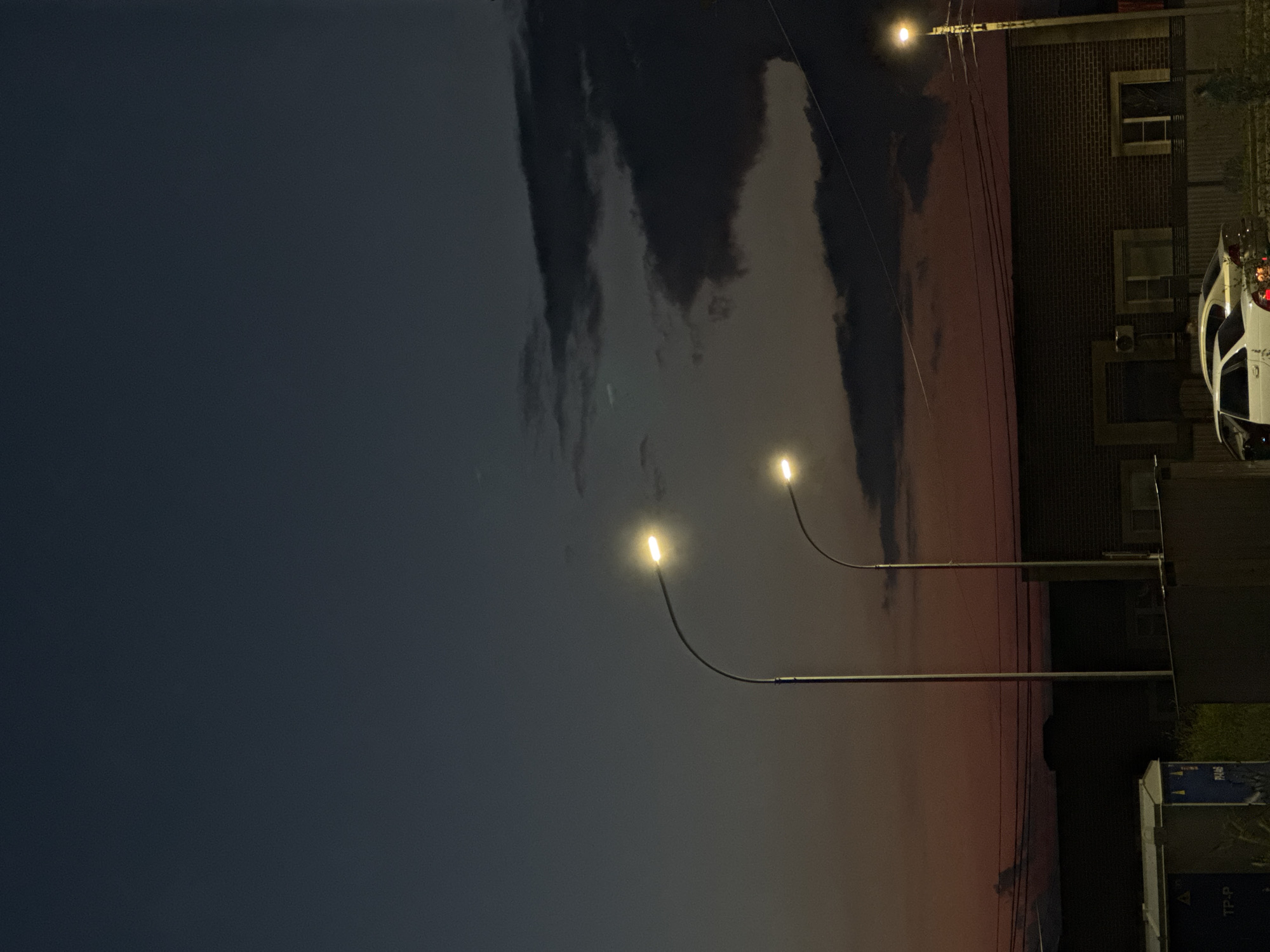 The image depicts a street scene at dusk in Mikhaylovsk, Russia. Two streetlights illuminate a brick building with several windows. Two cars are parked in front of the building, one white and one light-colored. The sky is a gradient of dark blue at the top, fading to orange and pink near the horizon, with dark clouds present. Power lines are visible stretching across the scene. In the foreground, there are some structures and vegetation.