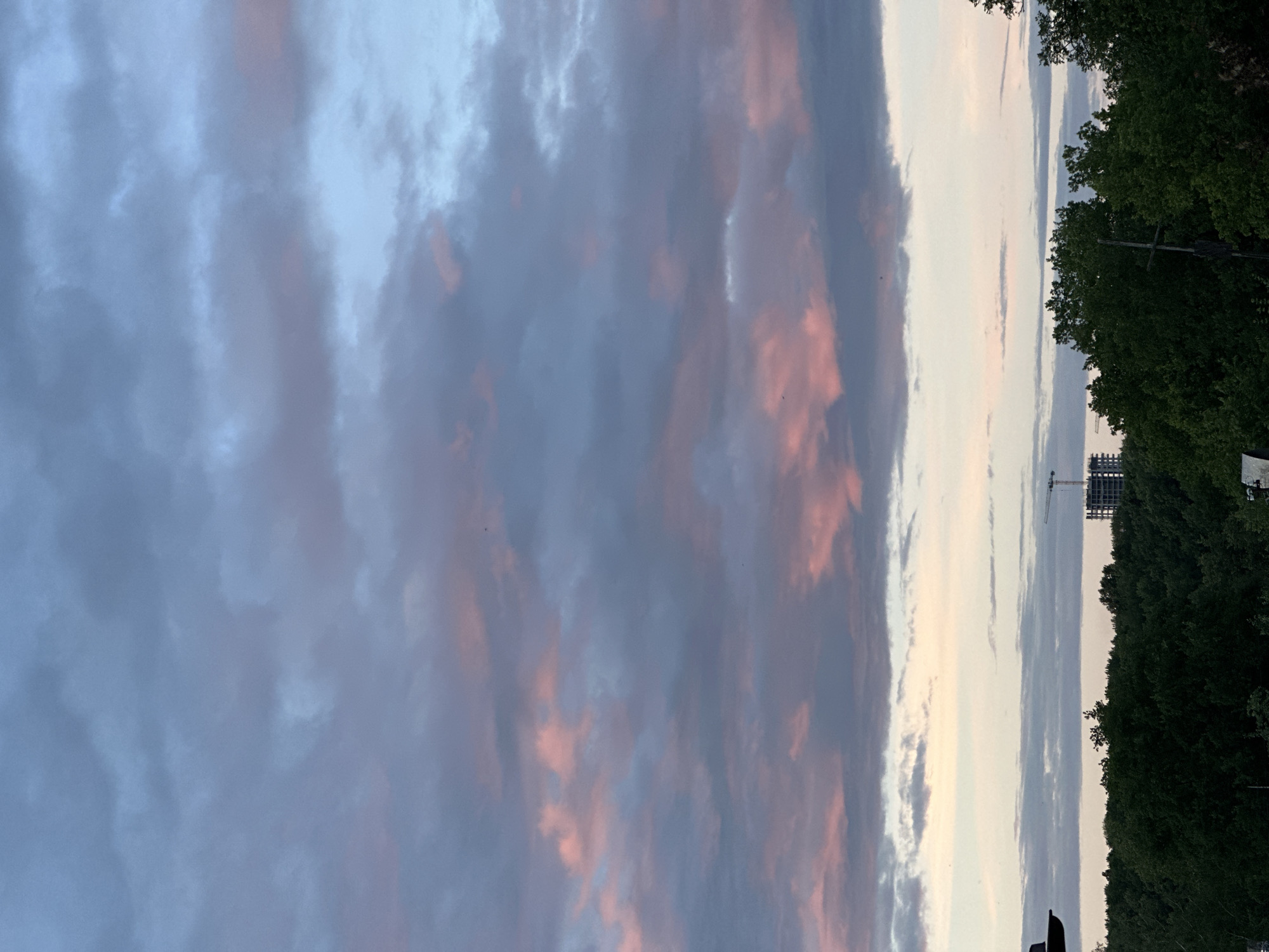 This is a landscape photograph taken at eye level, depicting a sky filled with dramatic clouds at dusk. The clouds are a mix of deep blues, grays, and soft pinks, suggesting the lingering colors of sunset. On the right side of the frame, a dense line of dark green trees forms a natural border. Beyond the trees, a construction site is visible with a prominent crane and a partially built structure. In the distance, a utility pole with crossbars is also present. The overall mood is serene and tranquil, enhanced by the soft light and the implied quiet of the evening. The time of day is clearly evening, as indicated by the sunset colors in the sky. There is no visible text in the image. The location context is Nizhny Novgorod, Russia.