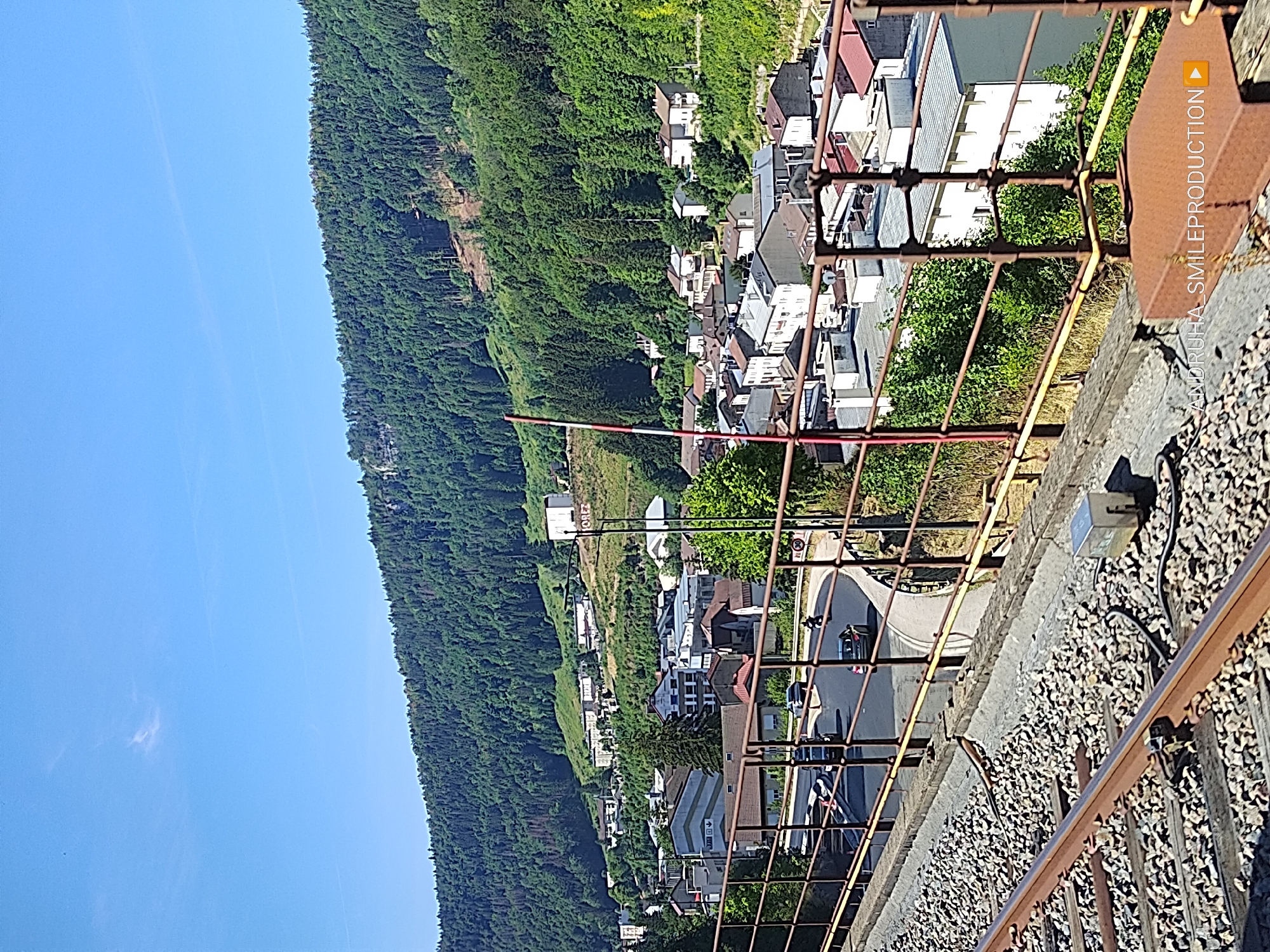 The image captures a view from an elevated railway track overlooking a town nestled at the base of a densely forested mountain in Hauts de Bienne, France. In the foreground, weathered train tracks covered in gravel occupy the bottom portion of the frame, with a rusty metal railing running parallel to them. Beyond the railing, a winding road curves through the town, with several vehicles, including cars and a motorcycle, visible on it. Buildings of various sizes and architectural styles, predominantly white with red or grey roofs, are clustered together, interspersed with greenery. The backdrop is dominated by a steep, tree-covered mountainside, with a clear blue sky above. The scene is bathed in bright sunlight, suggesting a daytime setting. There are no visible individuals in the image, but the presence of vehicles and buildings indicates human activity and habitation.