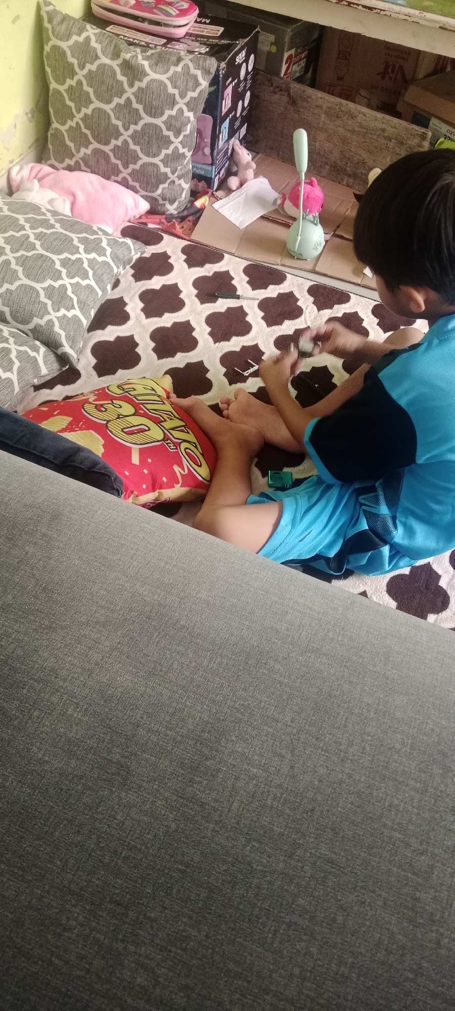 An overhead shot shows a young person sitting on a rug with a patterned design, playing with small white objects. The person is wearing blue shorts and a blue and black t-shirt, with their legs crossed and their feet visible. To their left, a red pillow with yellow lettering is visible. The rug is placed on a cardboard surface, surrounded by various items including cushions, boxes, and a small pink stuffed animal. A desk lamp with a light green base and a white stem is also present. The scene is set indoors, likely in a residential area.
