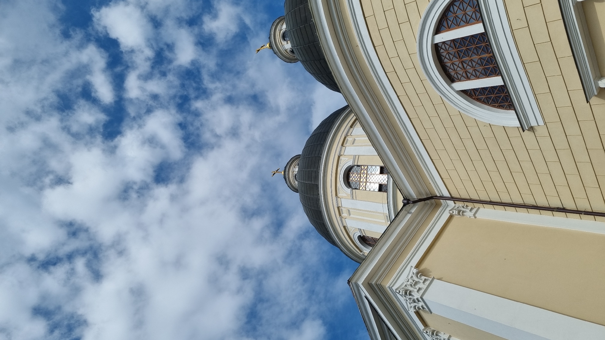 The image shows a low-angle view of the domes and facade of the Ascension Cathedral in Chernivtsi, Ukraine. The sky is blue with scattered white clouds. Two of the cathedral's dark domes are visible, each topped with a golden cross. The facade of the building is light yellow with white decorative elements, including pilasters and window frames. The windows are arched and have intricate lattice work. A dark downspout runs vertically along the facade. The architectural style suggests a religious building. There are no people or any discernible action taking place in the image.