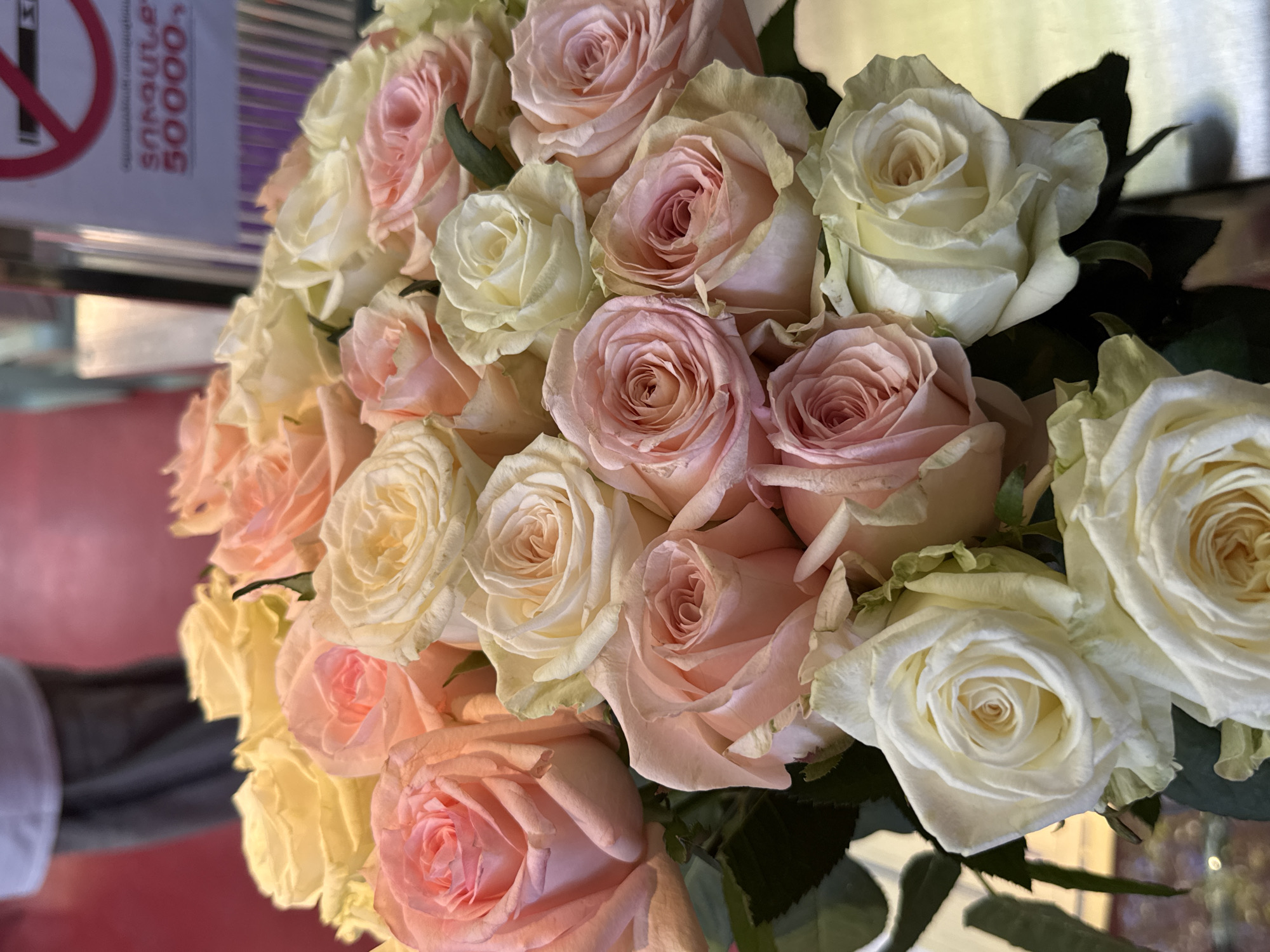 A close-up, high-angle shot presents a bouquet of roses in varying shades of pink and off-white. The roses are densely packed together, showcasing their delicate petals. Some green leaves are visible amidst the blossoms. A blurry gray sleeve on a pink blurred surface suggests that someone is holding the bouquet. To the left, out of focus, there is a white object with red circle and slash on it, along with some script.
The image was taken in Yerevan, Armenia.