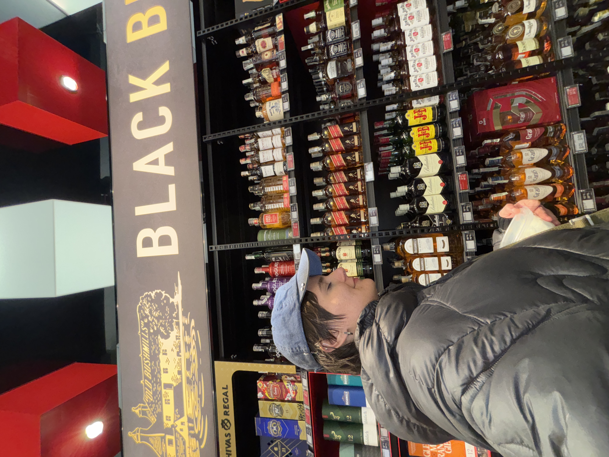 The image depicts an individual browsing the liquor aisle of a large supermarket in Sofia, Bulgaria. The setting features multi-tiered black metal shelving units extensively stocked with various brands of distilled spirits, including whiskies and bourbons such as Johnnie Walker (Red Label), Jack Daniel's, Jim Beam, Grant's, Bushmills, Chivas Regal, Ballantine's, Jameson, Dewar's, and Buffalo Trace. Price tags are visible on the shelves below the products.
Above the shelves, a prominent sign displays an illustration of a building and tree alongside the text "OLD BUSHMILLS" and a partial "BLACK B" in large white and yellow lettering, indicative of "Black Bushmills". A partial sign to the left also displays "CHIVAS REGAL". The ceiling above consists of alternating red and black rectangular panels, each fitted with circular recessed light fixtures.
A single individual is observed in profile, positioned on the left side of the frame and facing towards the right. This person, appearing to be a young adult, has short, dark hair and wears a light blue denim baseball cap reversed on their head. A small earring is visible on their left ear. They are dressed in a dark, likely black or dark grey, puffer jacket. Their right hand, partially visible, holds a small white container. The individual is focused on the products displayed on the shelves.