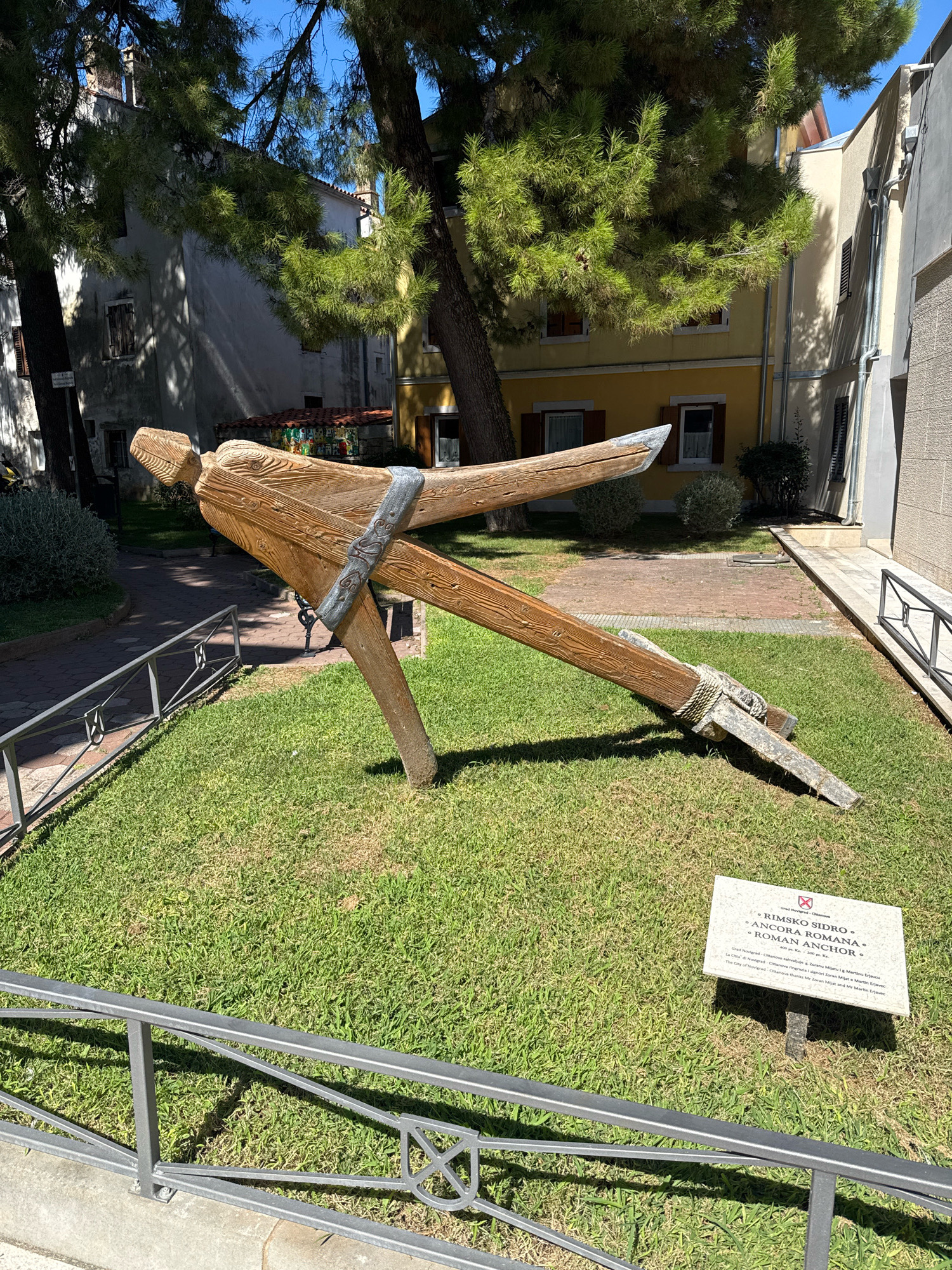 The image depicts an outdoor scene in Grad Novigrad, Croatia, featuring a large wooden sculpture of a Roman anchor displayed on a grassy area. The sculpture is positioned in the foreground and is the primary subject. It is made of weathered wood and metal, with visible rope details. Behind the sculpture, a paved pathway is visible, bordered by a metal railing. In the background, there are buildings with traditional Mediterranean architecture, characterized by their stucco facades and terracotta tiled roofs. Lush green trees with dense foliage provide shade, dappling the ground with sunlight. A white informational plaque is placed in the grass to the right of the anchor sculpture. The sky is a clear blue, indicating a sunny day.