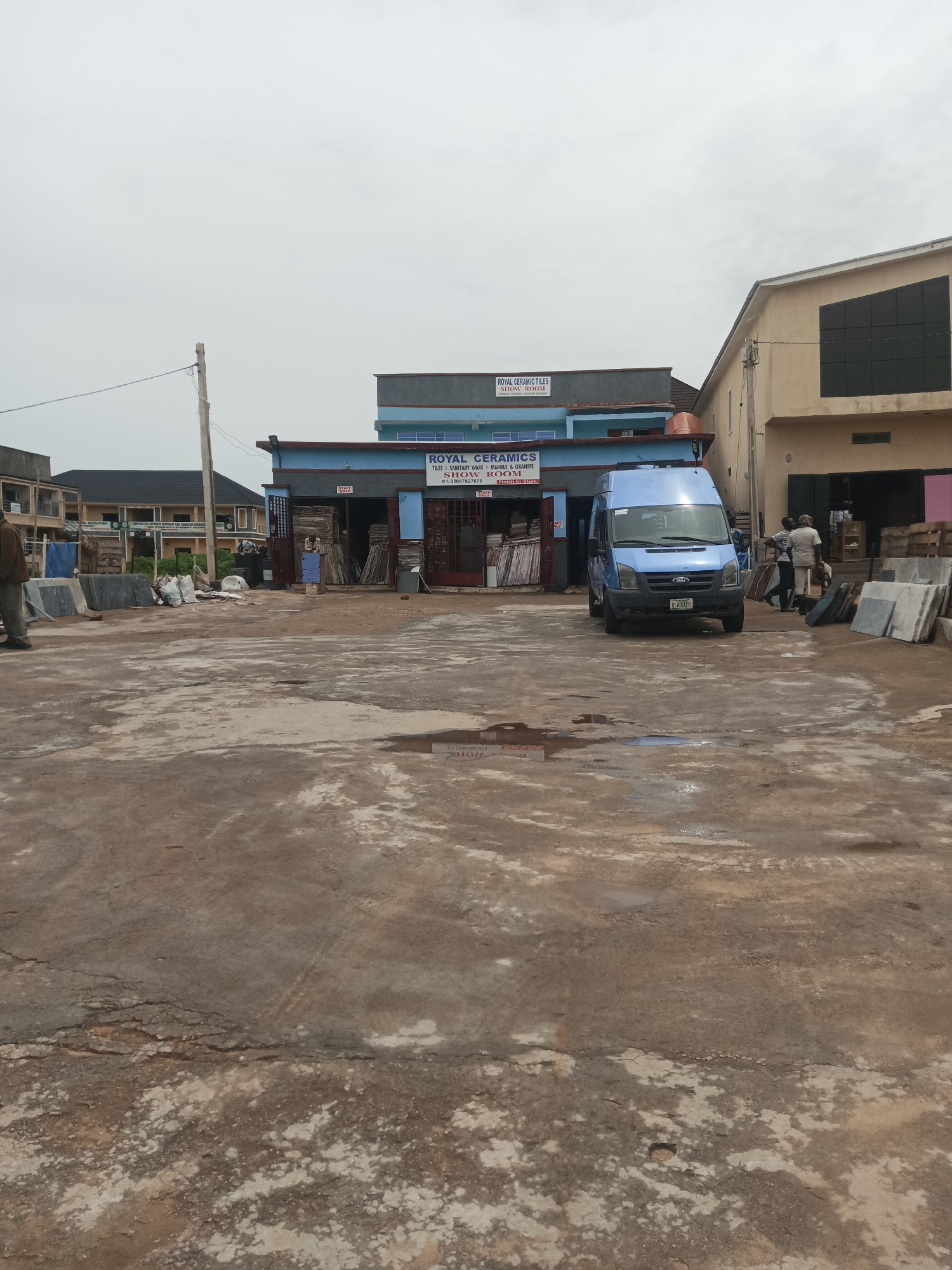 A scene unfolds in Gura Top, Nigeria, featuring a light blue van parked in a dusty, outdoor lot. The van, displaying a Nigerian license plate, is positioned in front of a building with blue and grey facades, where a sign reads "ROYAL CERAMICS SHOW ROOM." To the right of the van, several people are gathered, some carrying what appear to be slabs of material. In the foreground, the ground is uneven, with patches of dried mud and puddles, suggesting recent rainfall. Scattered around the lot are stacks of large stone or tile slabs. In the background, more buildings are visible, including a larger, beige structure with a dark, grid-like window on its upper section. The sky is overcast.