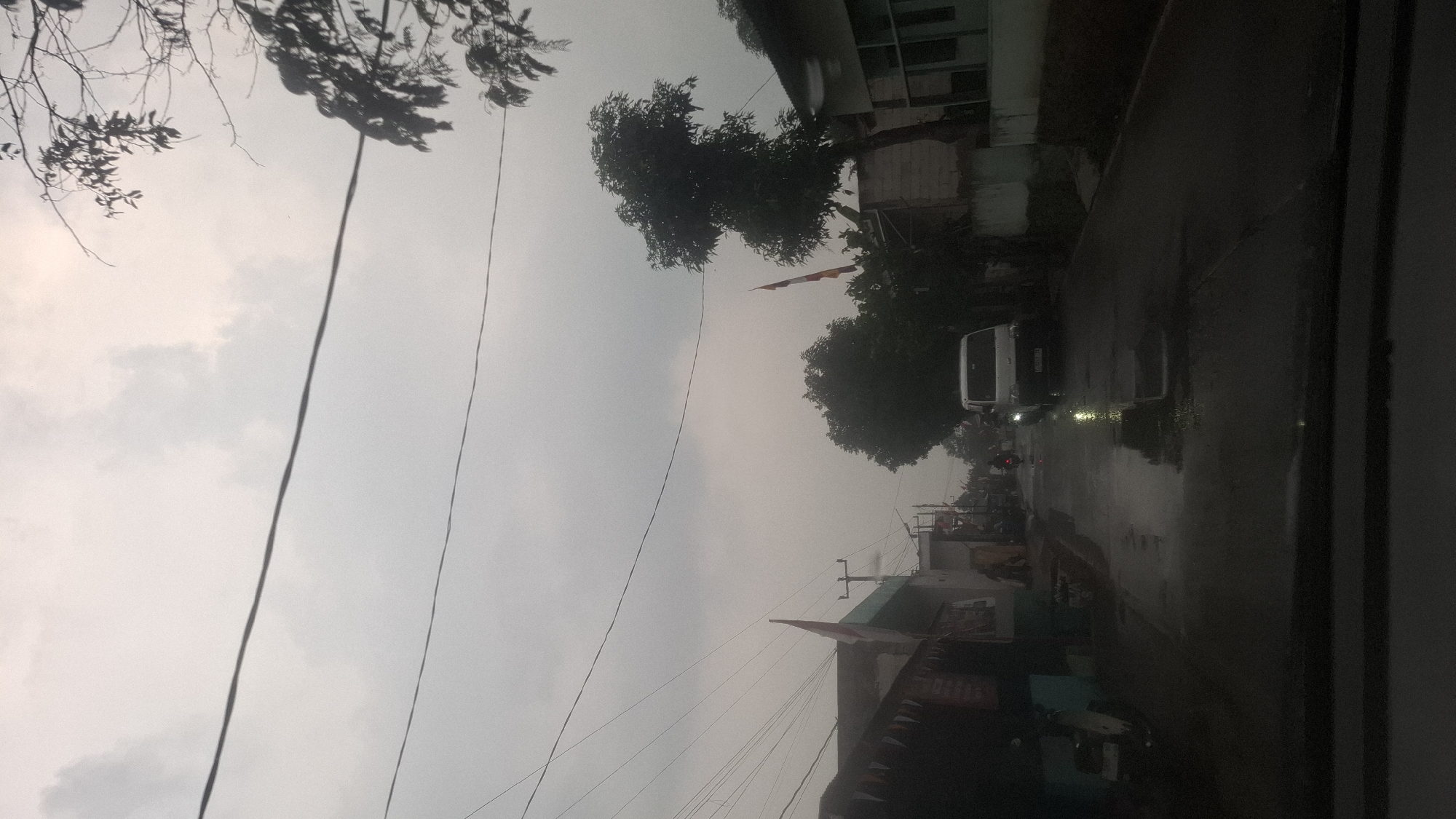 The image depicts a street scene in Cibinong Hilir, Indonesia, under an overcast sky. The road is wet, suggesting recent rain, with visible puddles reflecting the muted light. On the left side of the road, a dark-colored building with a turquoise awning is visible, alongside a parked white scooter. Further down the street, several other buildings and a power pole with multiple wires are present.
In the center of the street, a white van is driving away from the camera. To the right of the van, there's a lush green tree, and behind it, a building with a tiled roof. On the right side of the street, another building with a light-colored wall is partially visible. Flags, appearing to be Indonesian flags, are mounted on poles in front of some of the buildings, adding a touch of national identity to the scene. The overall atmosphere is subdued due to the cloudy weather.
