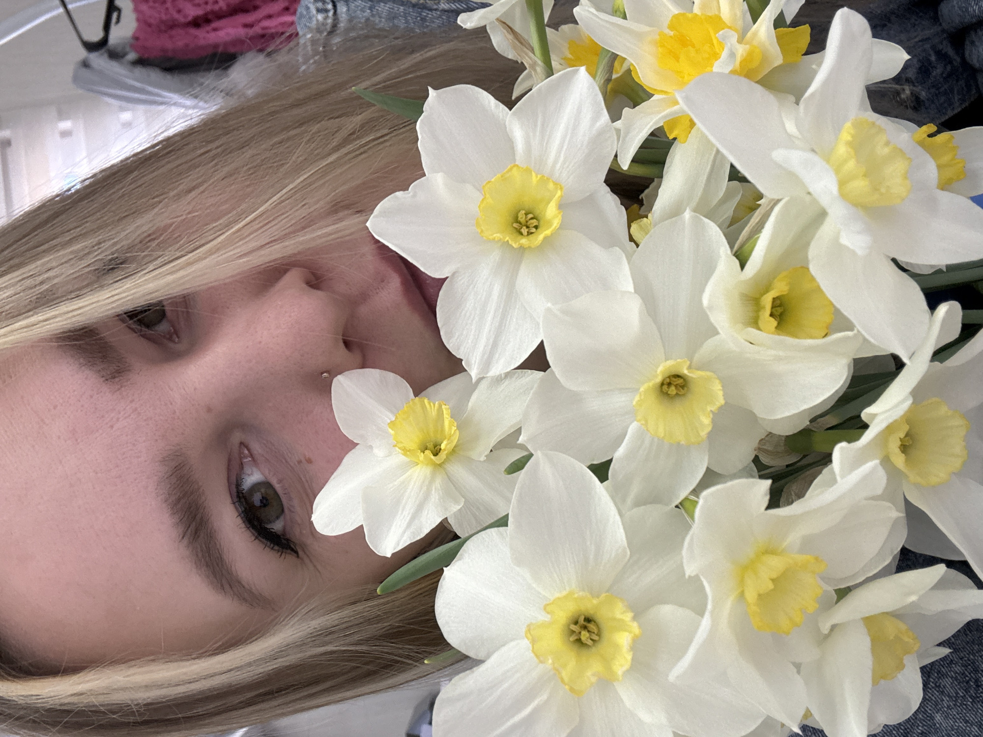 A close-up photograph depicts a woman with blonde hair, tilted to the left, partially obscured by a bouquet of white and yellow daffodils. Her left eye is visible with dark eyeliner. A silver stud is seen on her upper left lip. The background is blurry, revealing glimpses of a denim-like fabric and what appears to be a pink knitted item. The image does not provide any information to place the location in the city of Noviy, Russia.