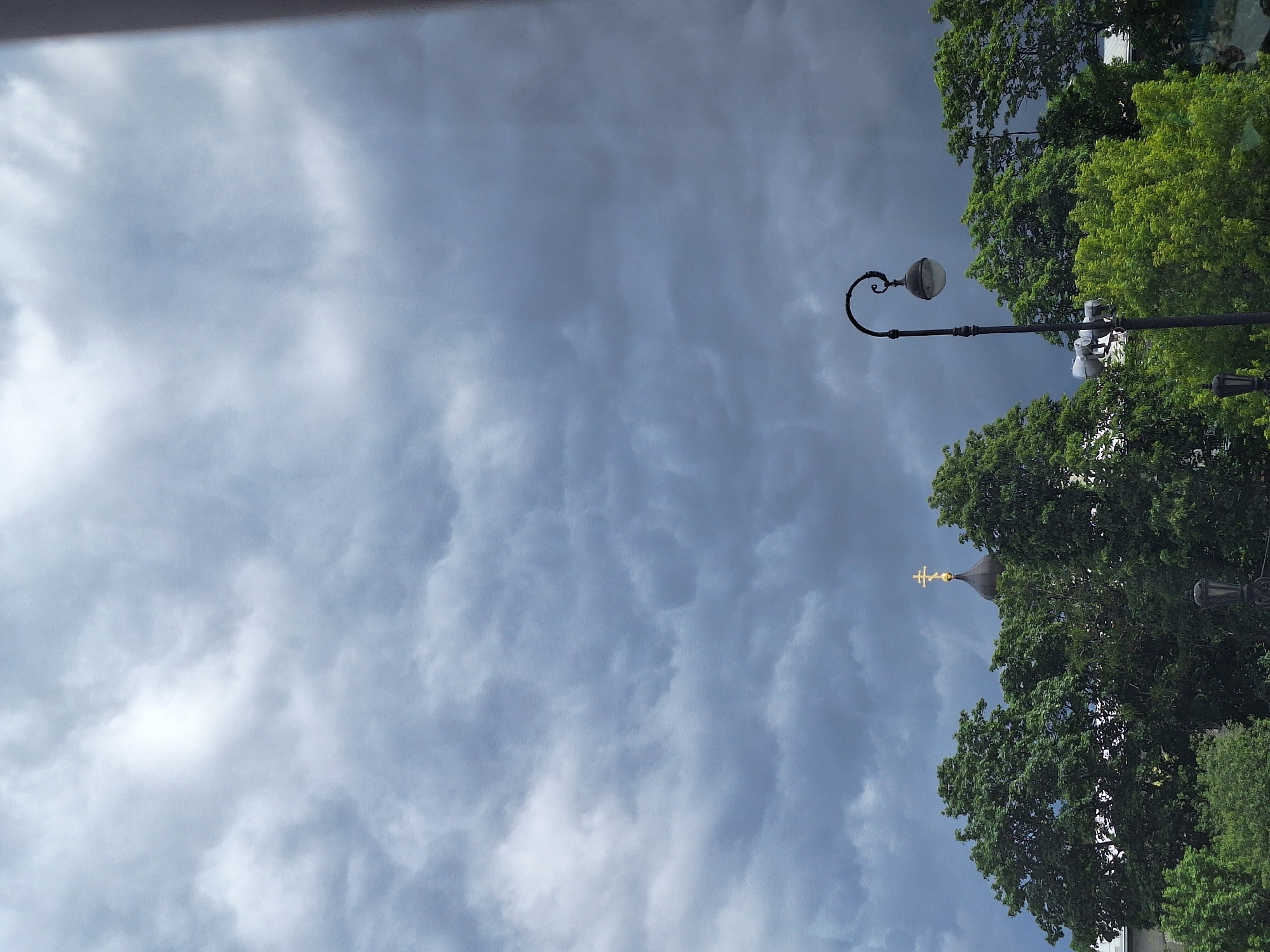 The image shows a view of the sky over Saint Petersburg, Russia. The sky is filled with dramatic, dark grey storm clouds, with some lighter patches of sky visible. In the foreground, the tops of lush green trees are visible, with the golden cross of a church dome peeking through the foliage. A vintage-style street lamp with a spherical light fixture stands on the right side of the frame. The overall impression is one of impending rain, with the city's architecture and greenery silhouetted against a turbulent sky.