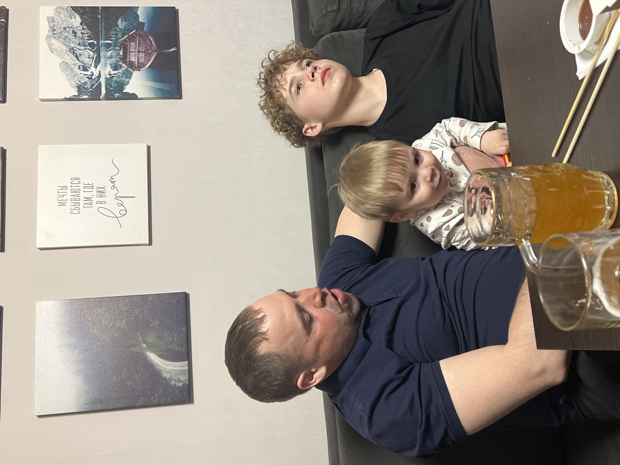 The image shows three males of varying ages relaxing on a dark sofa in what appears to be a casual indoor setting. An adult man with short dark hair and a beard, wearing a dark blue polo shirt, is lying or reclining, looking towards the right side of the frame with his mouth slightly ajar. Next to him, an older boy with curly light brown hair, dressed in a black long-sleeved shirt, is also lying back, gazing upwards. A baby or toddler with blonde hair, wearing a patterned light-colored onesie, is positioned between the man and the older boy, looking directly at the camera with a curious expression.
On a dark wooden table in the foreground, there are two glass mugs containing an amber-colored beverage, likely beer, with one appearing full and the other partially consumed. Beside the mugs, a small white bowl holds chopsticks, and a miniature white container holds a dark sauce, suggesting the consumption of an Asian meal like sushi.
The wall in the background is adorned with several framed canvases. The top left features two square canvases displaying a tranquil scene of mountains reflected in water, with a small boat. Below these, a white rectangular canvas shows black Cyrillic text that translates to "Dreams come true where they are believed in." A third rectangular canvas at the bottom left depicts a hazy, atmospheric landscape.
The overall scene suggests a relaxed family moment, possibly during an evening meal or gathering. The presence of Cyrillic text is consistent with the provided location context of Perm, Russia.