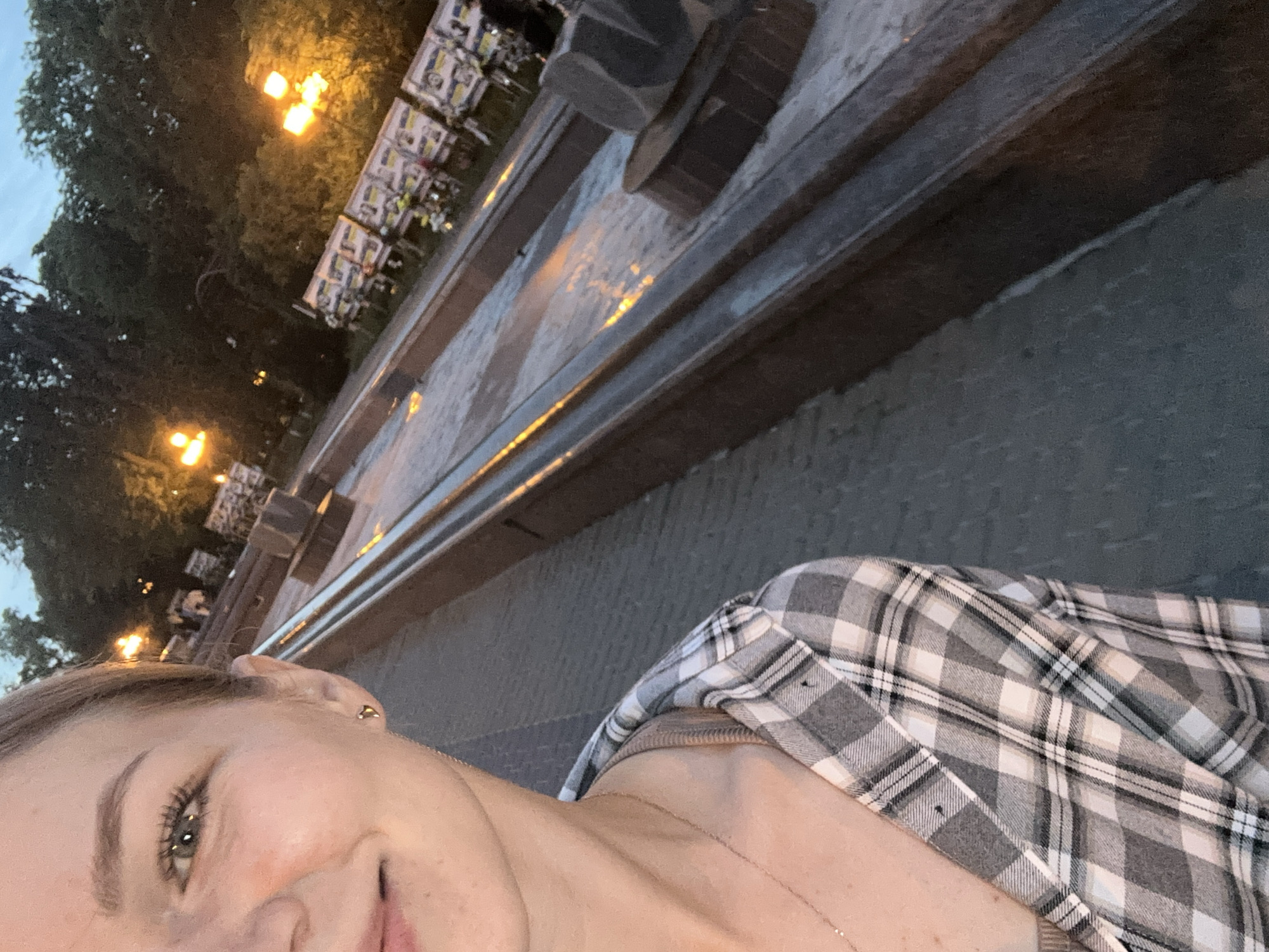 The image depicts a close-up, angled view of a woman on the left, with an urban park scene in the background at dusk or night.
The foreground features the upper left side of a woman's face and upper torso. Her right eye, with visible eyelashes and light-colored iris, is open and looking slightly to the right of the frame. Her right earlobe displays a small golden heart-shaped stud earring. Her skin appears fair, with a slight flush on the cheek. A subtle smile is present on her partially visible lips. She wears a plaid shirt in shades of grey, white, and black, open at the collar to reveal a light brown or beige undershirt. A delicate gold chain necklace with a small pendant hangs around her neck. Her hair, light brown or blonde, is pulled back and not fully visible.
The background shows a long, multi-tiered fountain or water feature, possibly a cascade, with dark, smooth stone or concrete edges. The water surface reflects the warm orange glow of distant streetlights. Beyond the fountain, rows of public benches are visible. Many benches are adorned with numerous small flags or banners, predominantly displaying the blue and yellow colors of the Ukrainian flag, suggesting a commemorative or patriotic display. Bright, spherical streetlights on tall poles emit an orange-yellow light, illuminating sections of the park. Dark, dense foliage of trees frames the upper background against a twilight blue sky.
The scene is located in Ternopil, Ukraine, specifically along the embankment of Ternopil Pond, likely near the **Central Embankment Cascade Fountain**, a prominent public space. The presence of Ukrainian national symbols on the benches indicates a public display of patriotism or remembrance, common in Ukrainian cities.