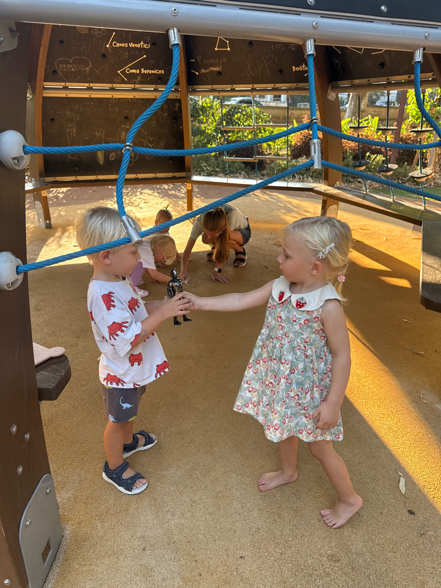 Two young Caucasian children, a boy and a girl, are interacting at a playground. The boy, on the left, has blonde hair and is wearing a white t-shirt with red crab graphics and grey shorts with a dinosaur print. He is wearing dark blue sandals. He is holding a small black action figure. The girl, on the right, also has blonde hair and is wearing a light-colored dress with a strawberry print and a ruffled collar. She is barefoot. They appear to be exchanging the action figure. In the background, another young child and an adult woman are visible, also at the playground. The playground has a sandy surface and a structure with ropes and wooden panels. The environment suggests an outdoor setting with natural light. The image does not provide specific information about the location being Limassol, Cyprus.
