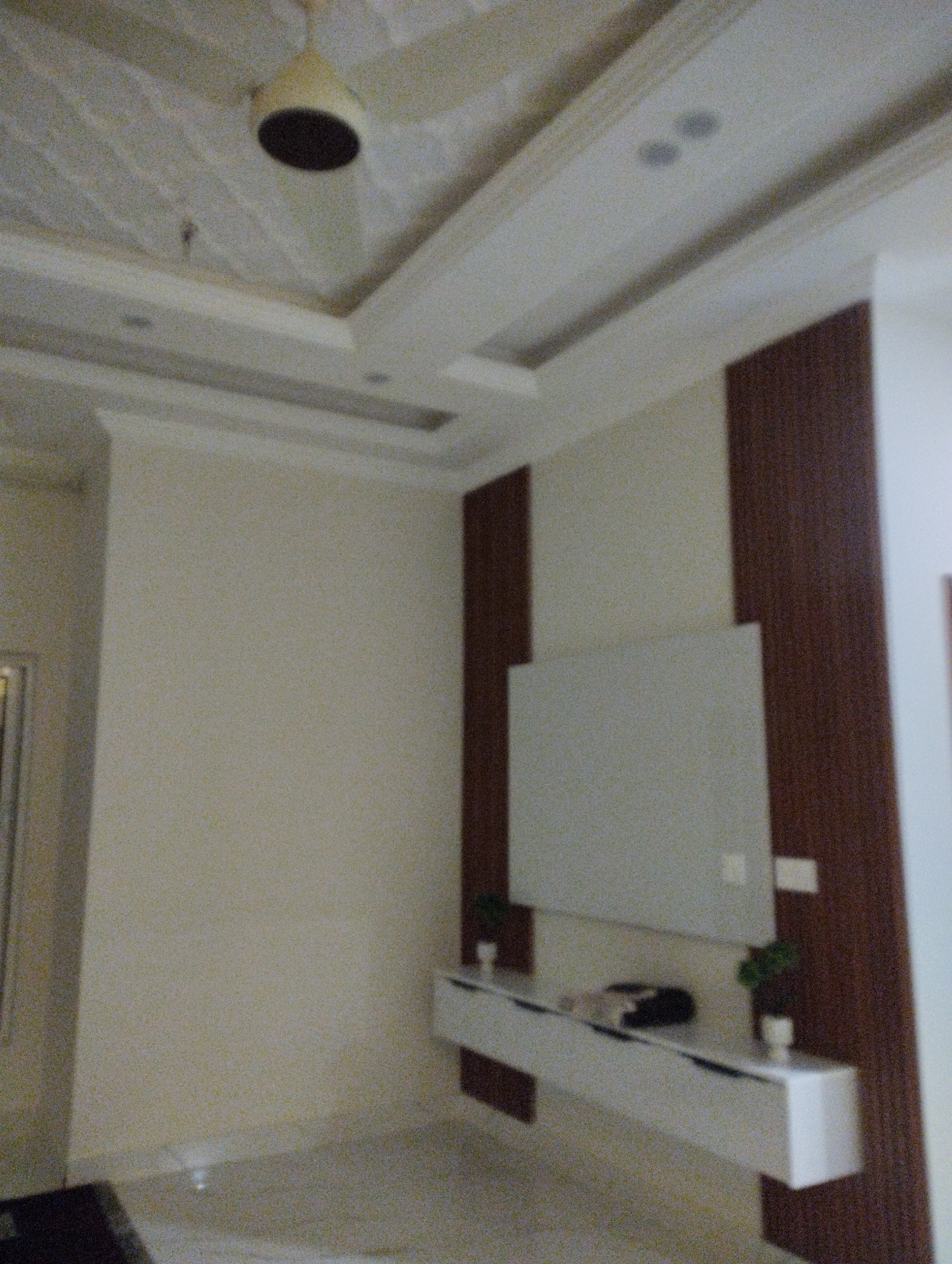 The image depicts an interior view of a room, likely a living room or entertainment area, in an unspecified location. The scene is dominated by a wall-mounted television and a floating media console. The television is a large, rectangular screen, appearing to be off. Below it, a sleek, white media console is suspended from the wall, featuring a minimalist design. Two small potted plants with green foliage are placed on the console, one on either side of a dark object that may be a soundbar or other electronic device. The wall behind the television is divided into sections, with a large, light-colored panel in the center and darker wooden panels on either side. The room features a textured ceiling with recessed lighting and a ceiling fan, the latter visible in the upper left portion of the image. The walls are painted in a neutral, light tone. The floor appears to be a light-colored tile or polished concrete. The overall aesthetic suggests a modern and contemporary design. There are no individuals present in the image.