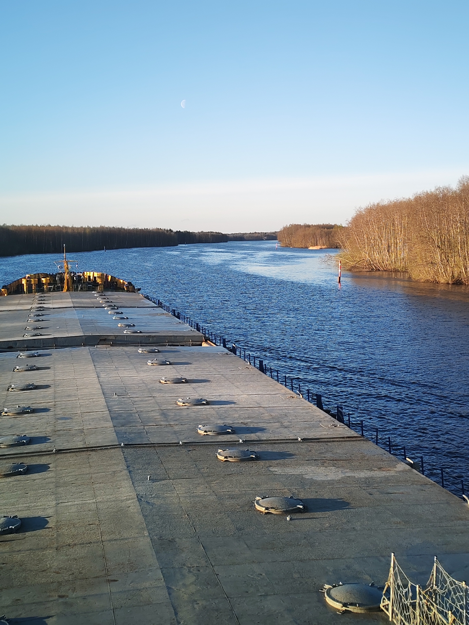 The image shows a river view from the deck of a barge or ship. The deck is flat, made of concrete squares with evenly spaced round hatches. A low metal railing runs along the edge of the deck. The river is wide, with tree-lined banks on both sides. A red and white buoy marks the channel. The sky is pale blue with a crescent moon visible. The location is Kurdiug, Russia.