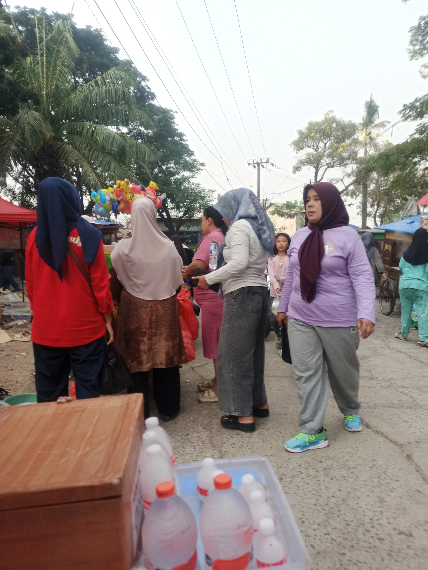 The image depicts an outdoor scene in Telagamurni, Indonesia. In the foreground, a wooden box and several plastic bottles filled with a pinkish liquid are visible on a gray, speckled surface. In the midground, several individuals are present. A woman in a purple hijab and a purple long-sleeved shirt stands facing right, wearing gray pants and blue and green sneakers. To her left, a woman wearing a gray hijab and a gray long-sleeved top, with dark gray pants, is also present. A child in a light pink top stands behind her. Further left, a woman in a red long-sleeved shirt and dark pants, with a blue hijab, is seen from the back, and another woman in a beige hijab and a brown, patterned skirt. In the background, a palm tree and other foliage are visible, along with some overhead wires. A cart with a blue canopy and another cart with a red canopy are also present, suggesting a market or roadside stall setting. Several colorful balloons are displayed on a string. The overall atmosphere appears casual, possibly a marketplace.