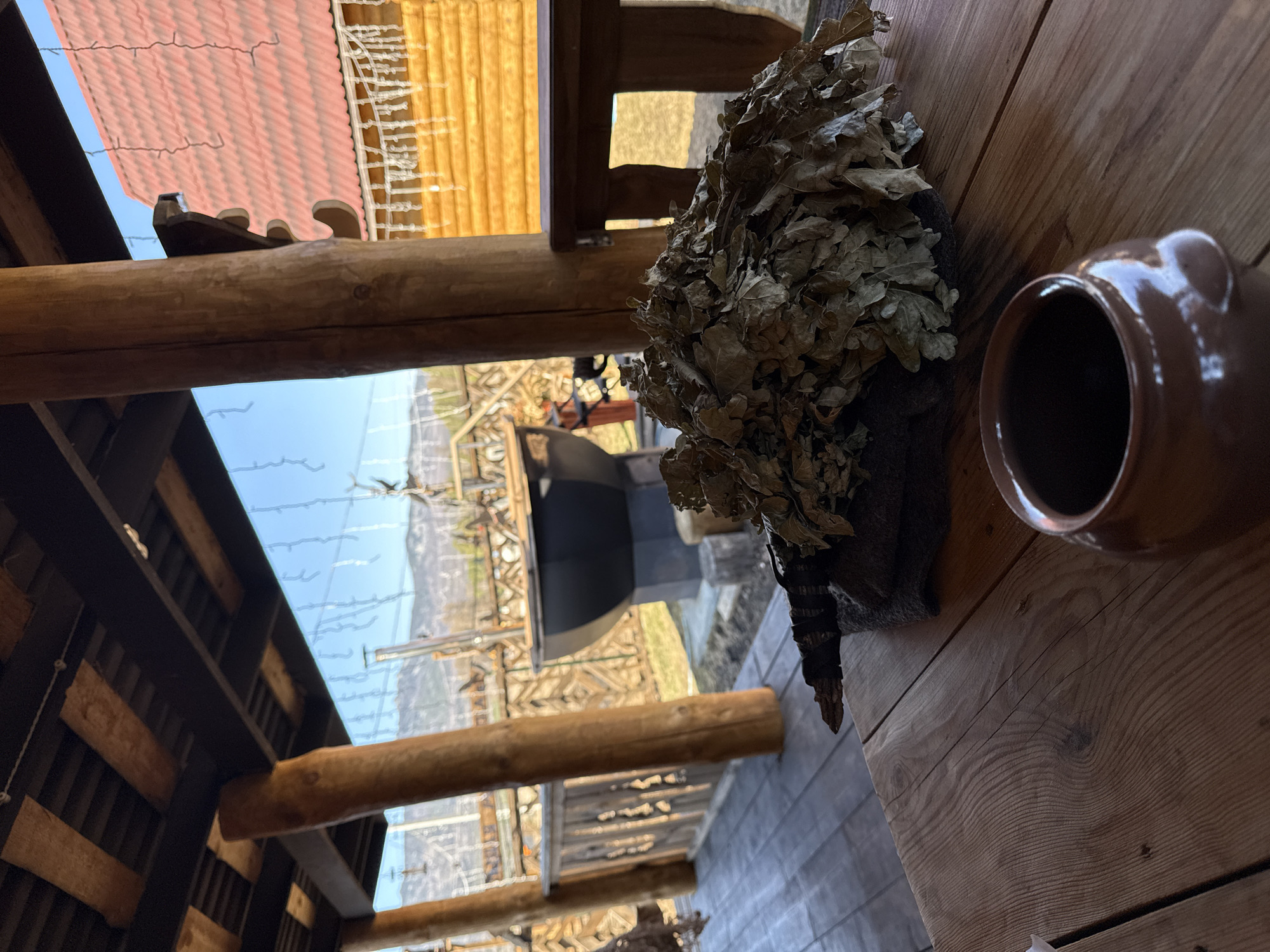 The image shows a scene inside a wooden structure, possibly a sauna or bathhouse, in Tyulyuk, Russia. A bundle of dried birch branches (likely a "venik" for use in the sauna) rests on a wooden table next to a brown ceramic pot. Through the structure's open side, a view of the surrounding landscape is visible, including metal structures, possibly related to the bathhouse or a nearby area. The roof above is visible, consisting of red tiles and decorative wooden elements. The overall lighting suggests it is daytime.