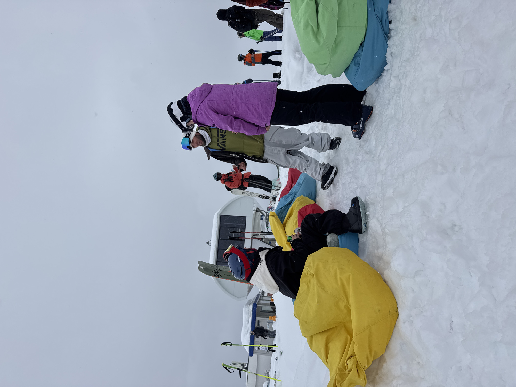 The image depicts a snowy outdoor scene, likely at a ski resort, in an unidentified location. There are several people present, some standing and some sitting on what appear to be colorful beanbag chairs in the snow.
In the foreground, a person is sitting on a yellow beanbag chair, wearing a blue helmet and a white jacket with a black torso. Ski equipment is nearby. Behind this person, there are other colorful beanbag chairs, including red and blue ones.
In the midground, a group of people are standing. One person is wearing a purple jacket and black pants, standing beside someone in a green "VANS" hoodie and grey sweatpants. Further back, other people are seen wearing ski gear, including orange jackets.
In the background, there is a building that appears to be part of the ski resort infrastructure, likely a ski lift station. Ski poles and a yellow barrier are visible in front of the building. The sky is overcast.